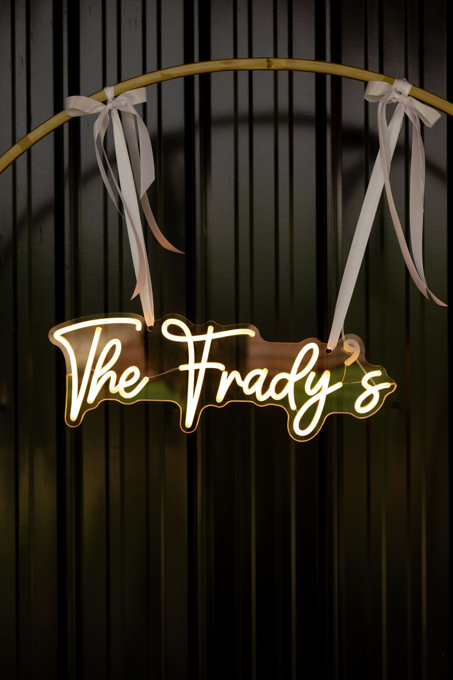Glowing neon sign reading The Frady's hung with white ribbon bows on a bamboo arch against dark backdrop.