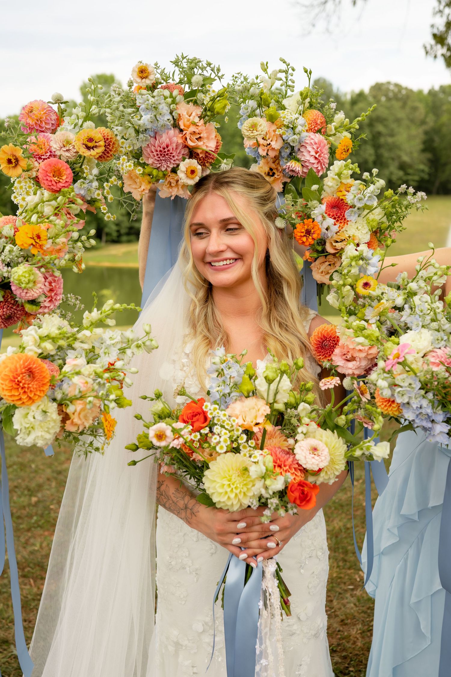 Radiant bride smiles holding colorful bouquet framed by lush floral arch with dahlias at outdoor garden wedding.