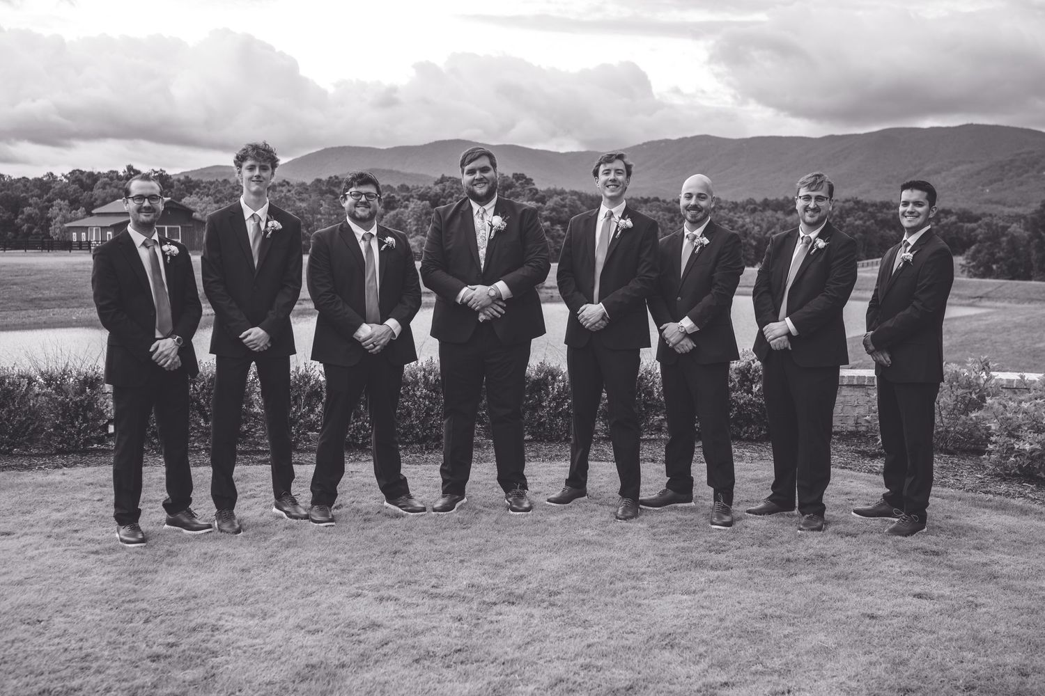Black and white photo of a groom and groomsmen in suits posing outdoors with mountain scenery at a wedding.