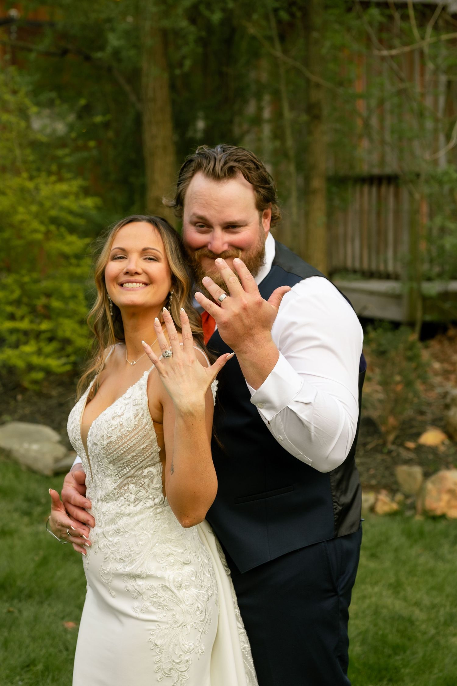 Newlyweds smiling and showing off wedding rings outdoors, bride in white gown and groom in navy vest.