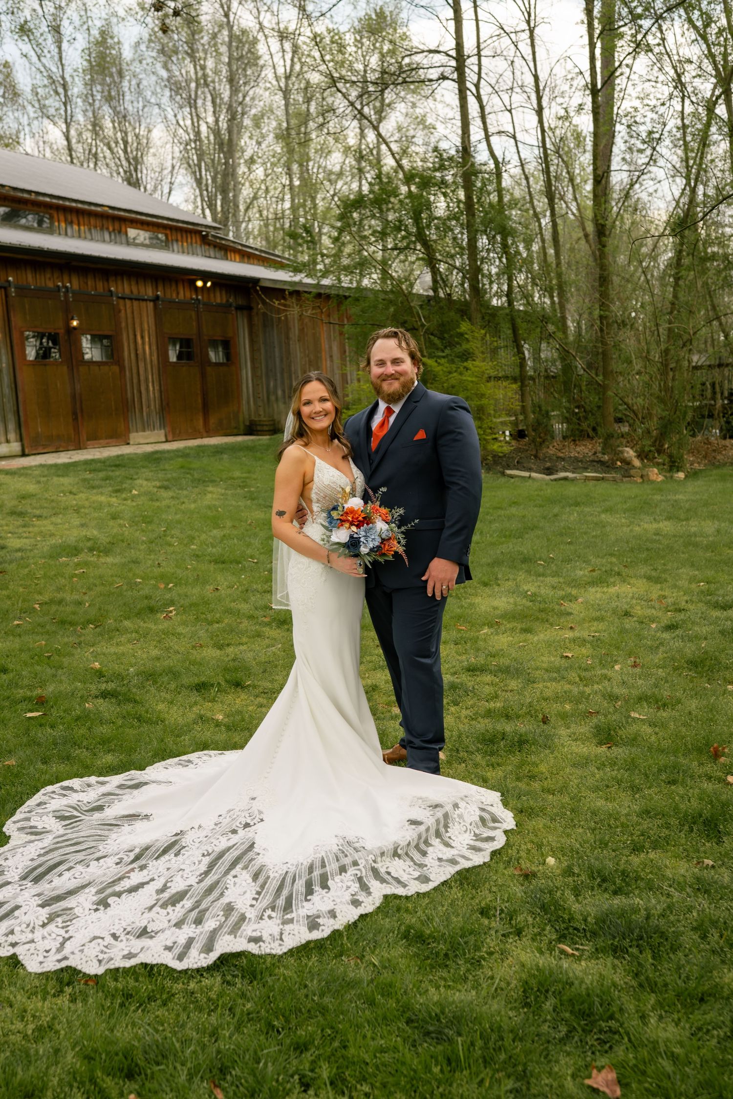 Bride and groom posing on green lawn, she wears a lace train gown holding a colorful bouquet.