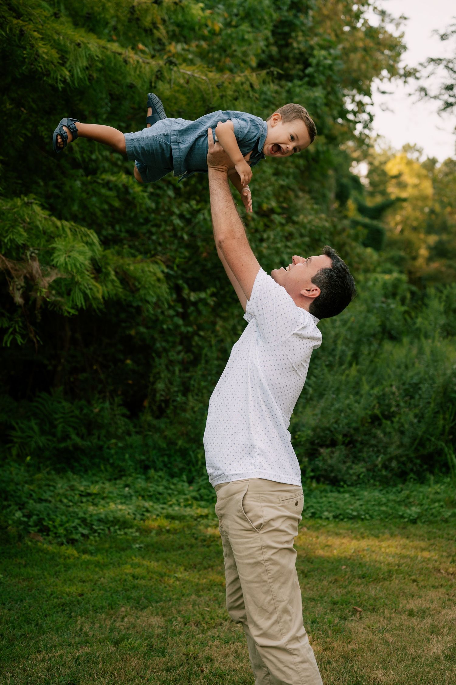 A person in a white shirt and khaki pants performs an expressive dance movement in a lush green outdoor setting.