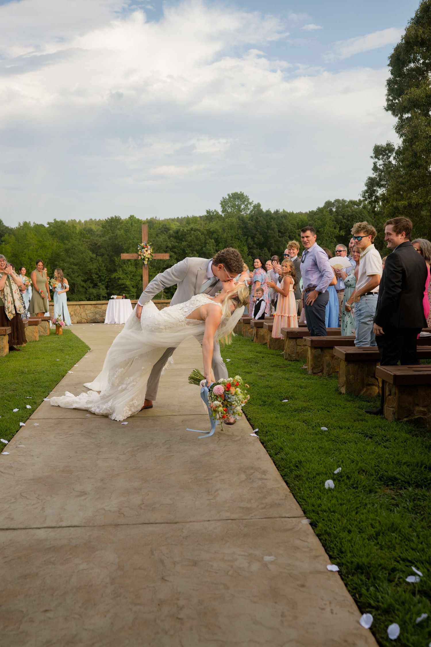 Bride and groom share a romantic dip kiss at outdoor wedding ceremony while guests celebrate along the aisle.
