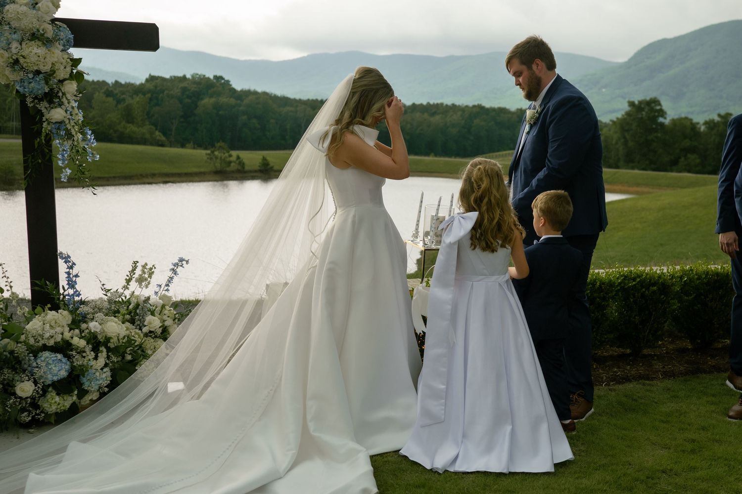 Bride and groom exchange vows outdoors with mountain views, flower girl nearby.
