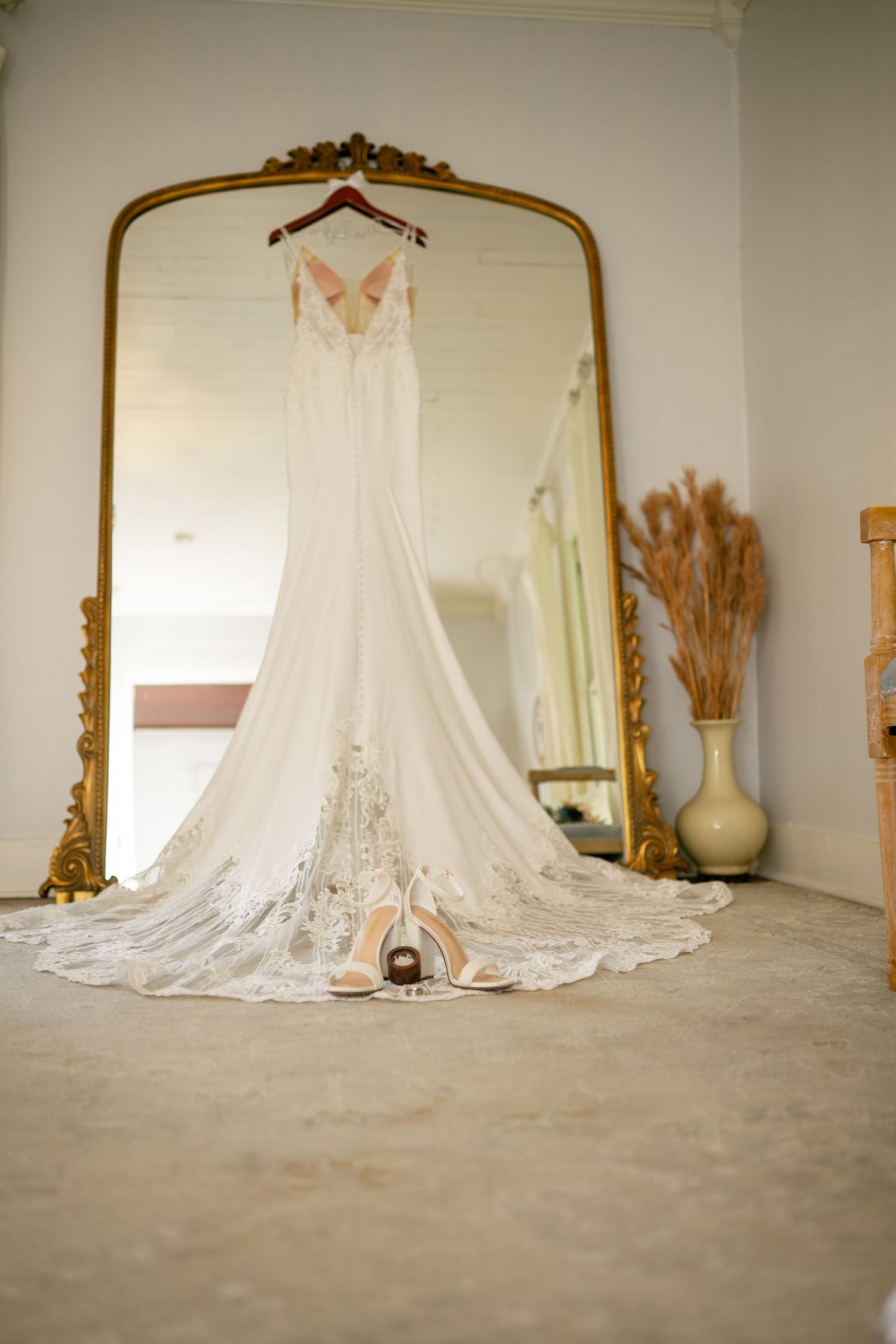 Elegant white lace wedding dress hanging on ornate gold mirror with bridal heels displayed on floor.