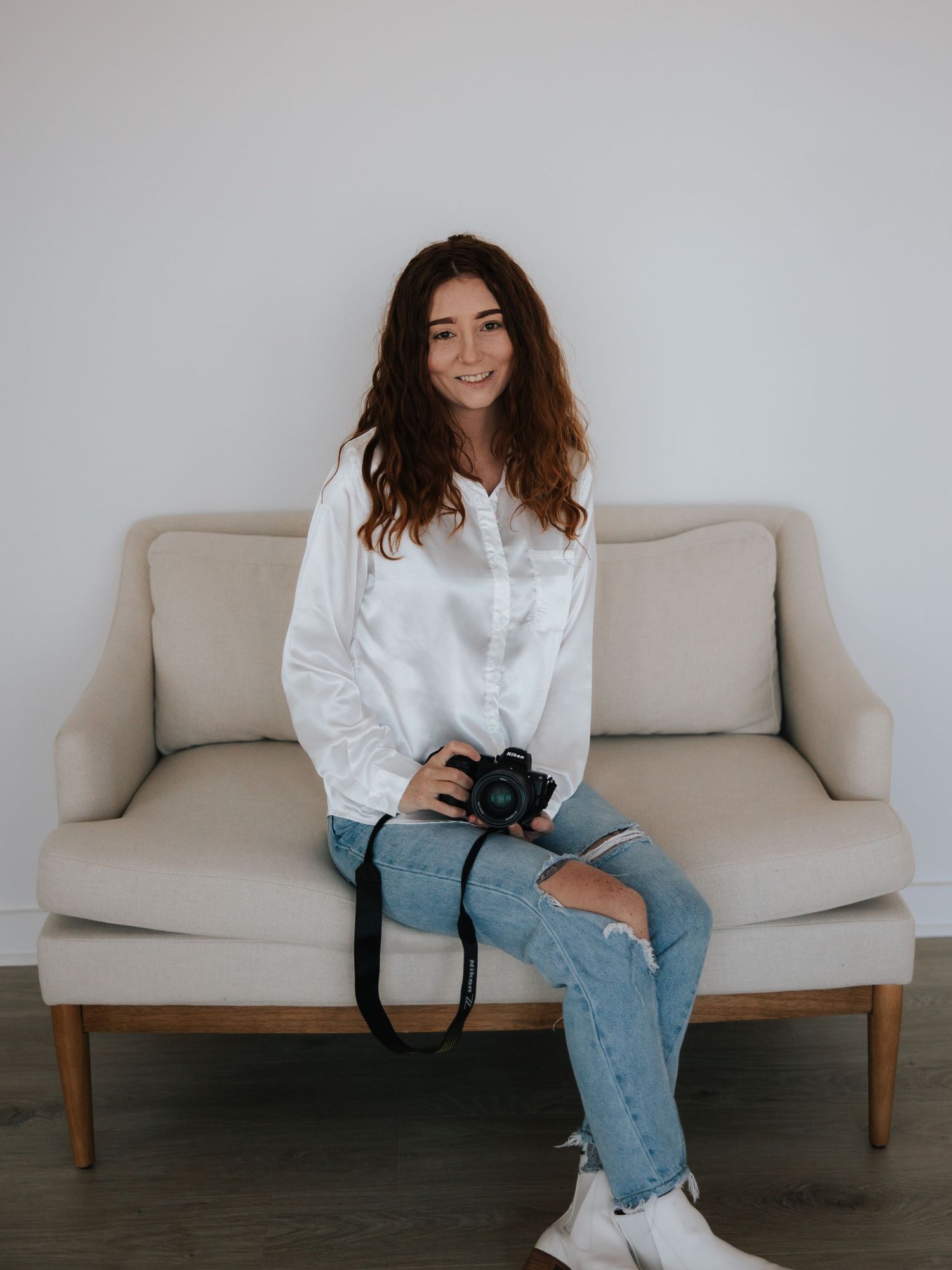 Photographer in white button down shirt and ripped jeans sits casually on beige couch holding professional camera.