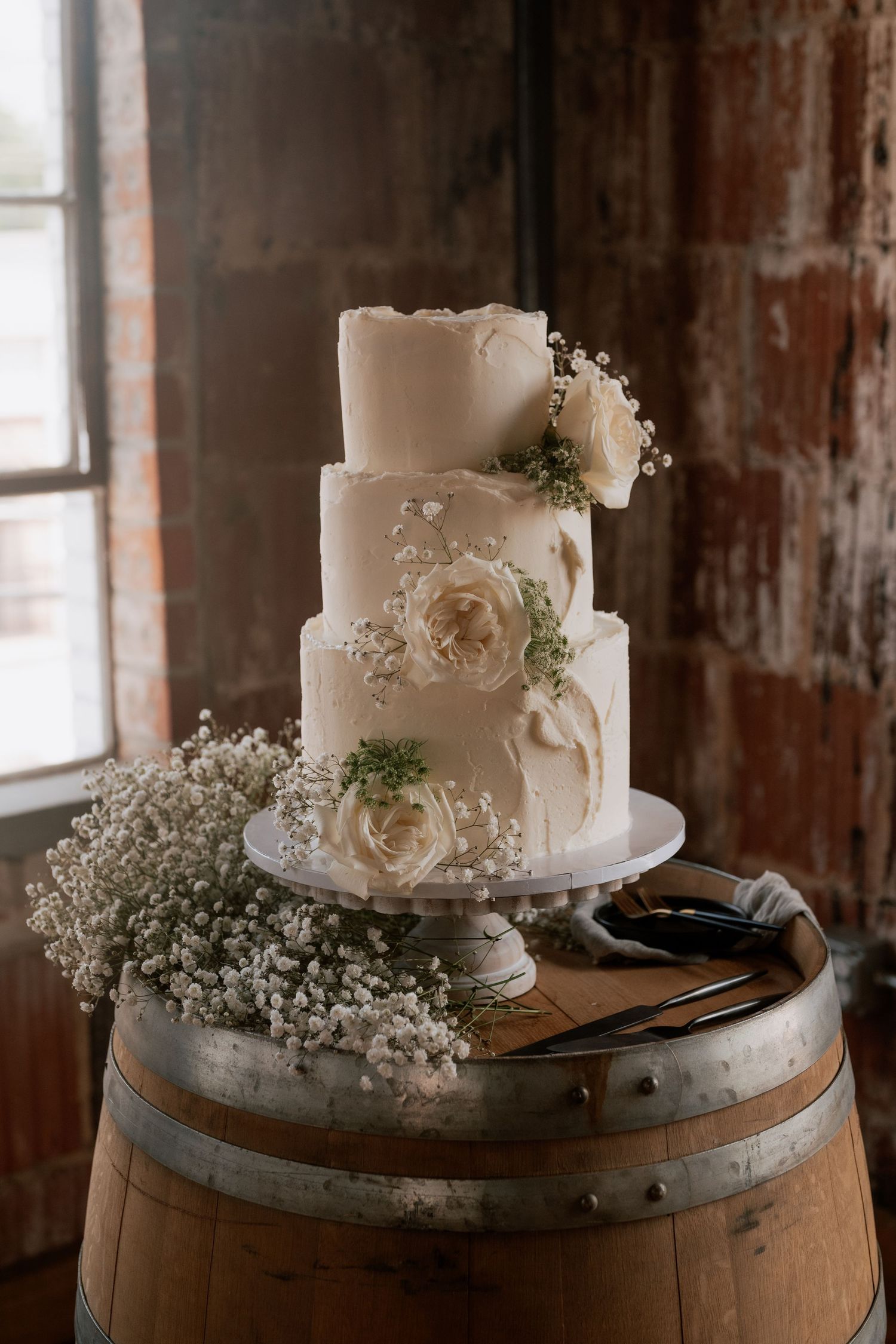Elegant white wedding cake decorated with roses and baby's breath sits atop rustic wooden barrel in vintage setting.