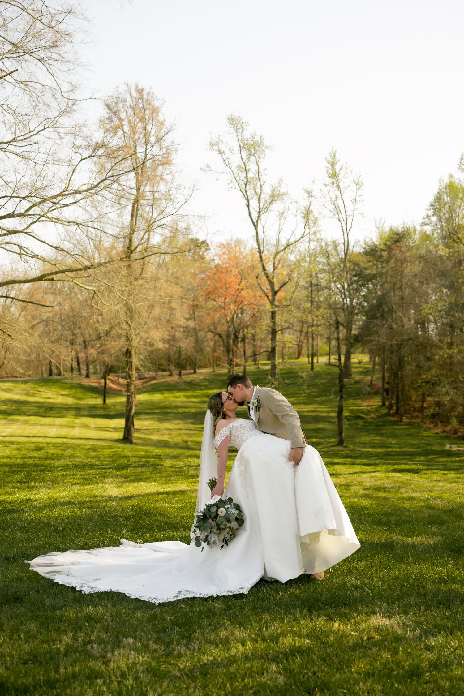 A groom dips his bride for a kiss on a lush green lawn surrounded by autumn trees in an outdoor wedding portrait.