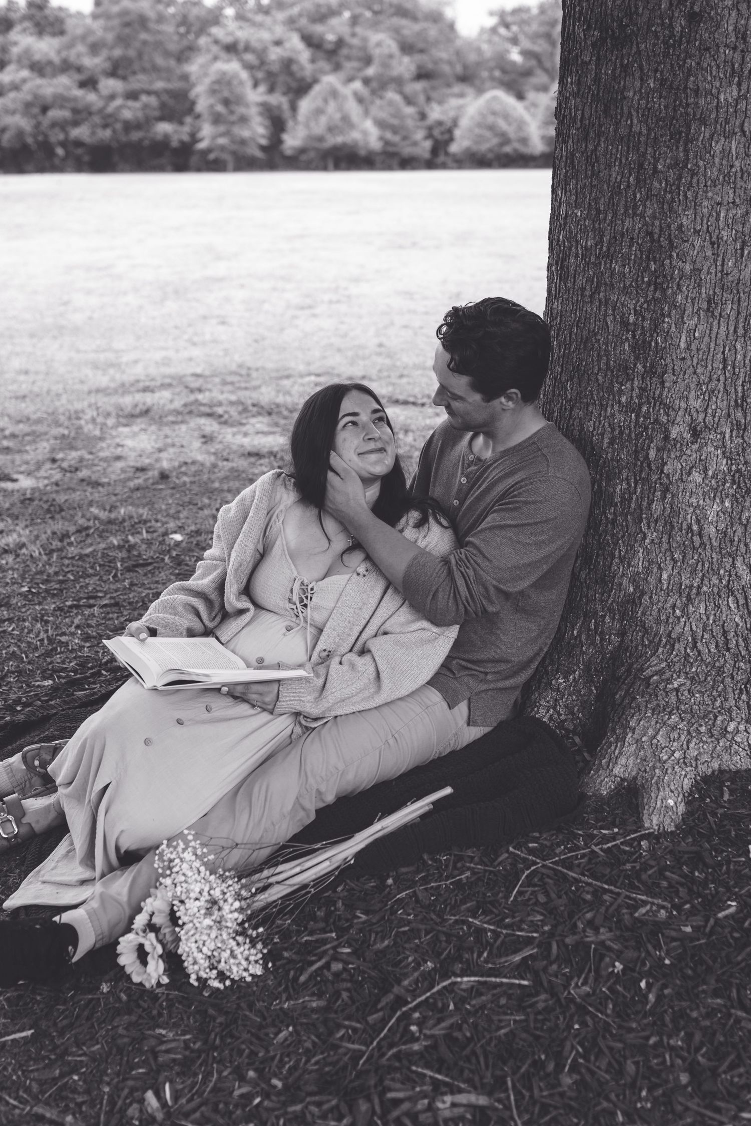 Black and white photo of a romantic couple sitting under a tree in a park, sharing a book and flowers.