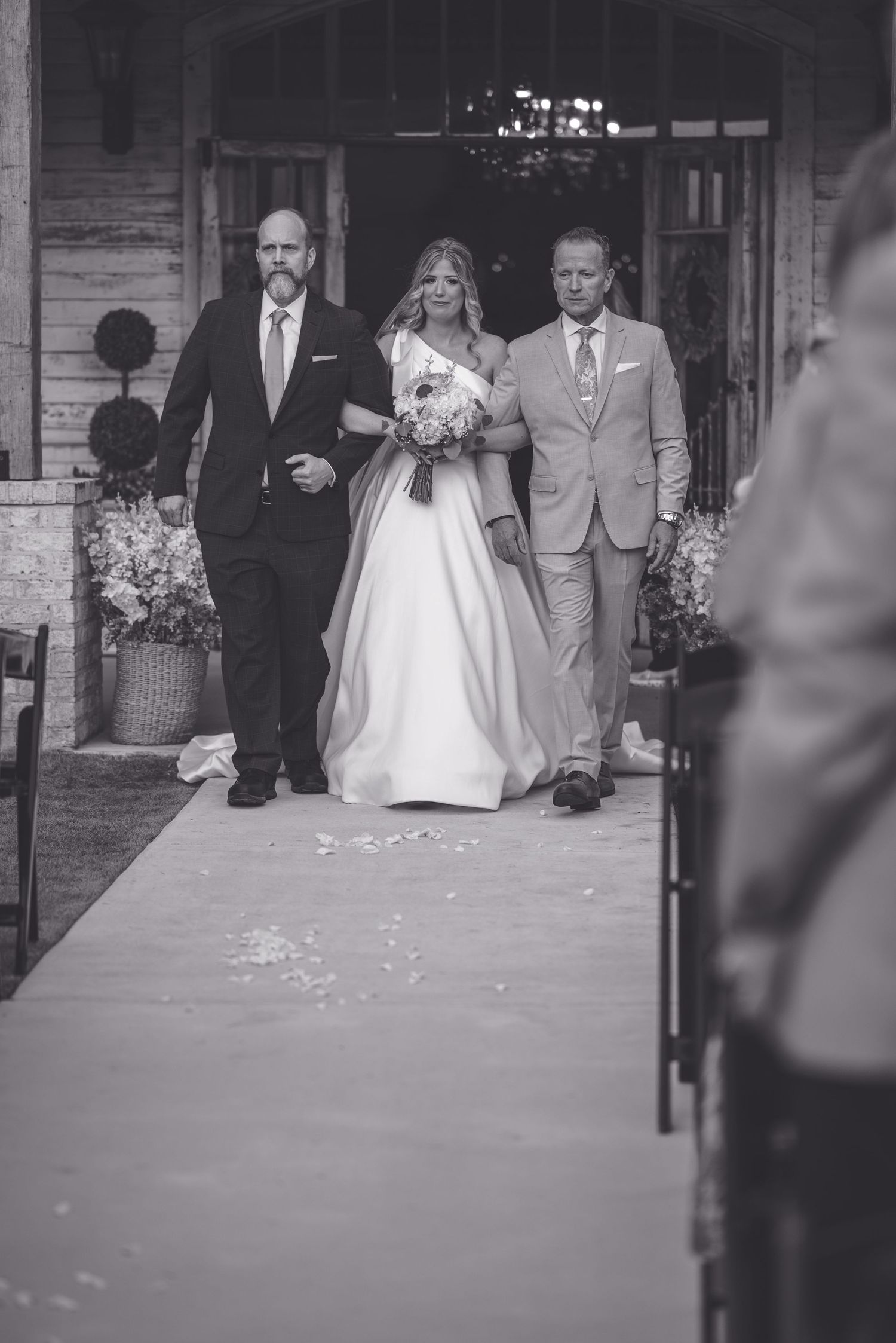 Black and white photo of bride in elegant gown being escorted down the aisle by two men at outdoor wedding ceremony.