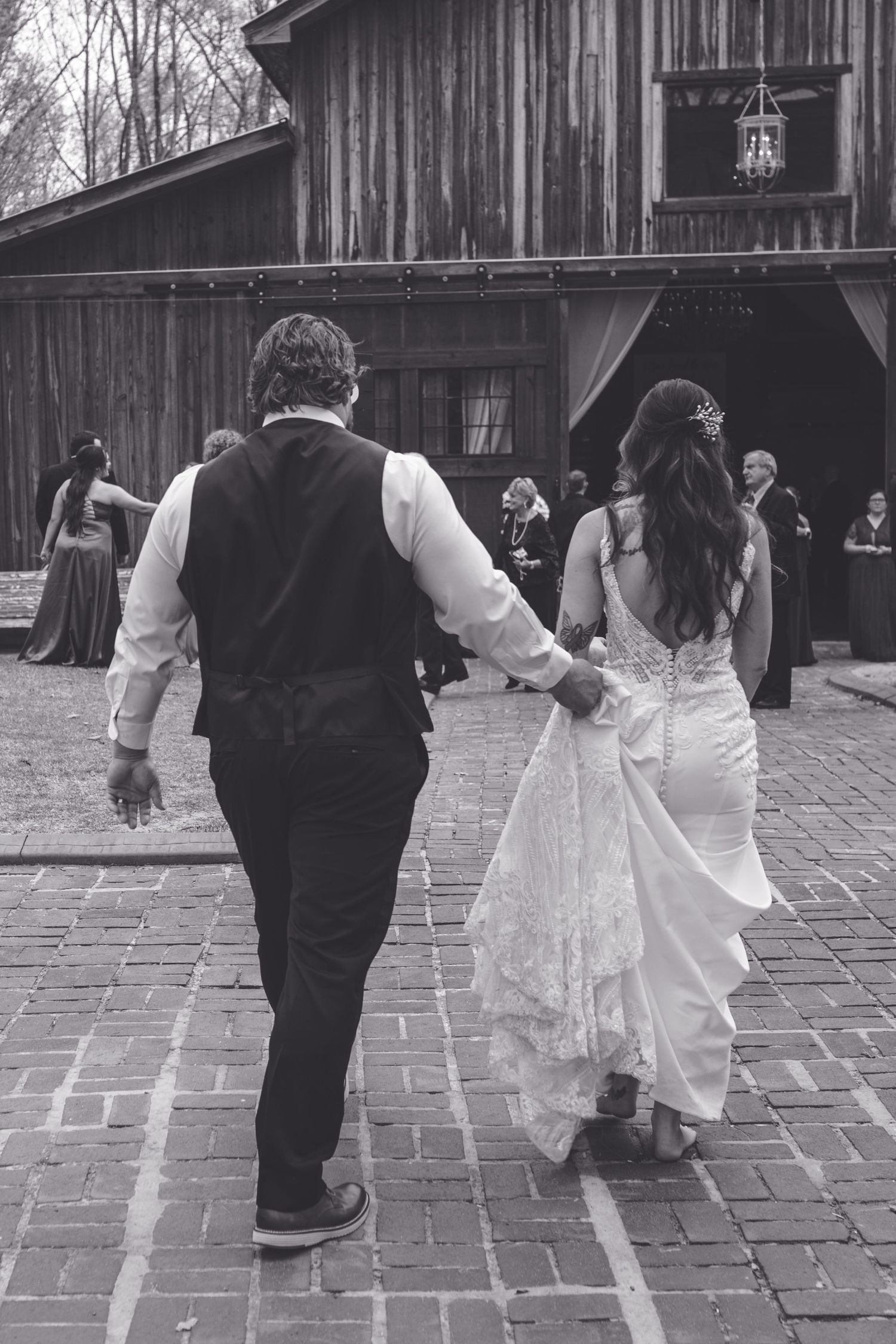 Black and white photo of bride and groom holding hands walking toward rustic barn wedding venue.