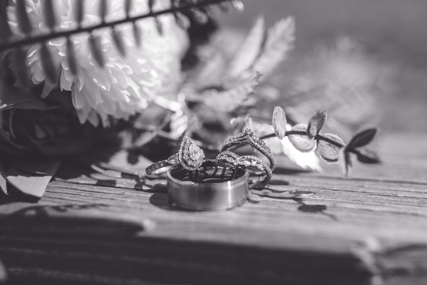 Black and white photo of elegant wedding rings and diamond engagement ring on wooden surface with floral bouquet backdrop.
