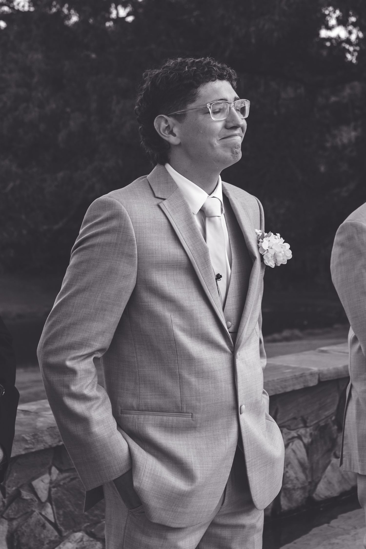 Groomsman in gray suit with boutonniere smiling at outdoor wedding ceremony.