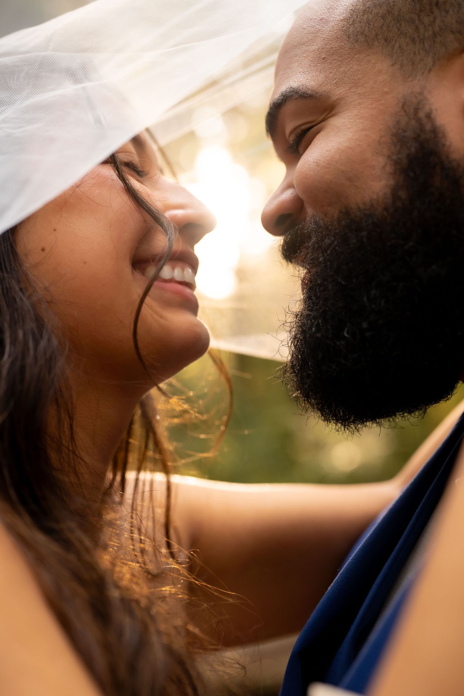 A smiling bride and bearded groom share a romantic moment under a wedding veil in soft golden sunlight.