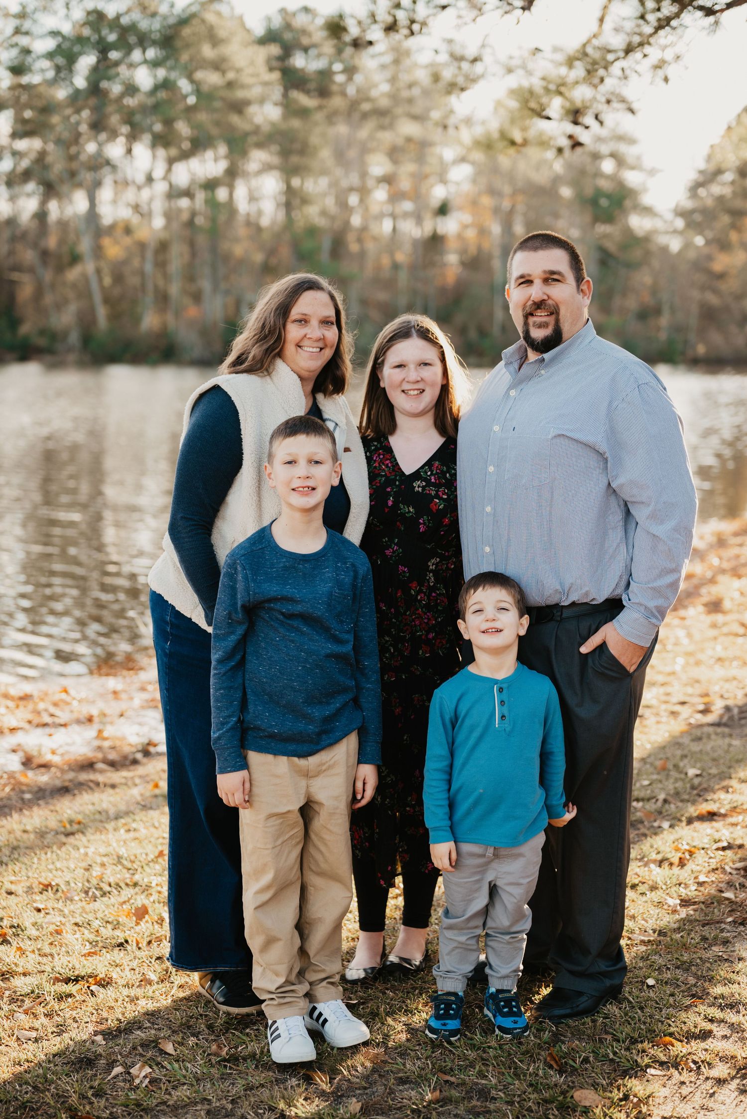 Happy family of five posing together by lakeside with fall trees in background.