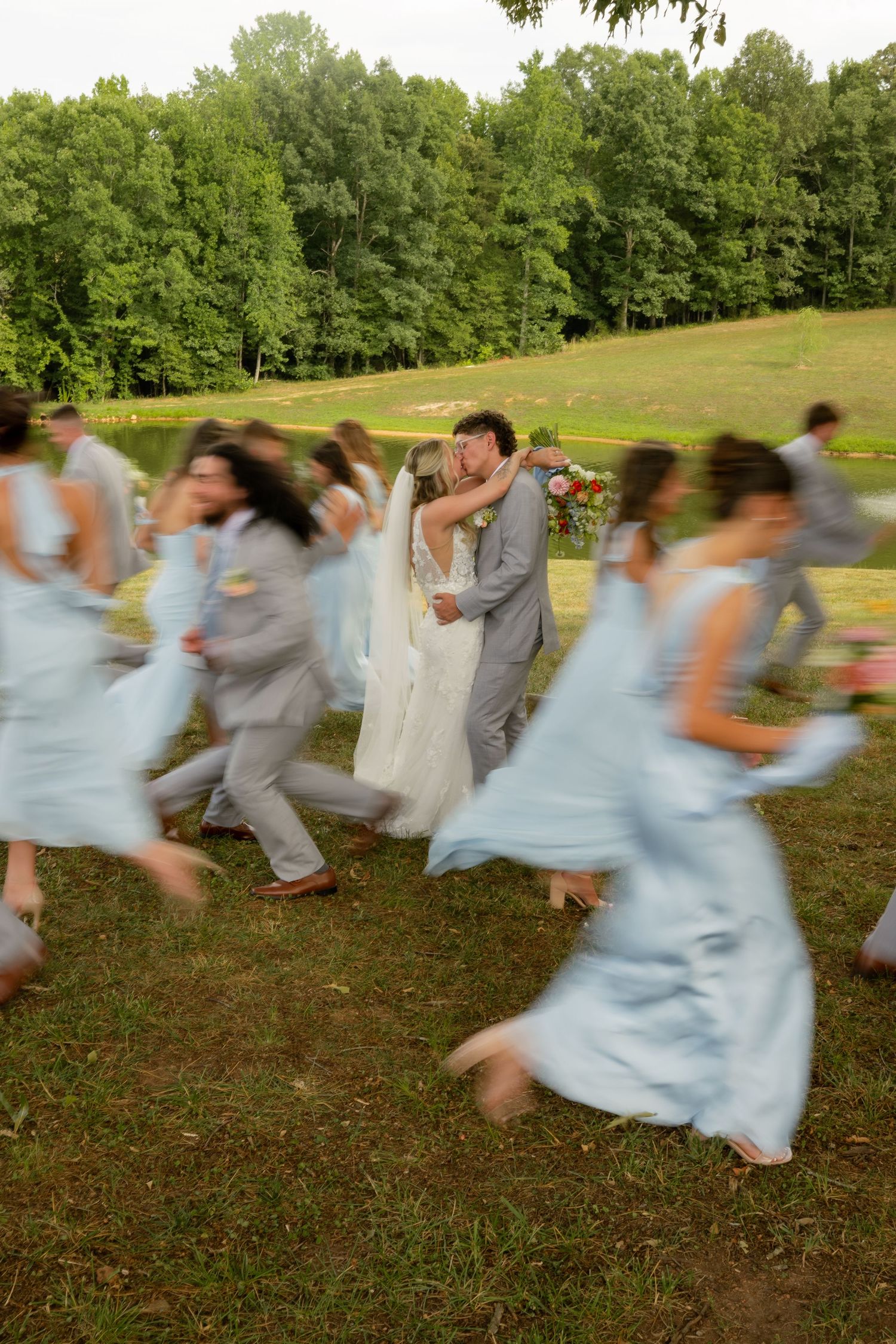 Wedding party surrounds kissing bride and groom in blurred motion on green lawn at outdoor summer wedding.