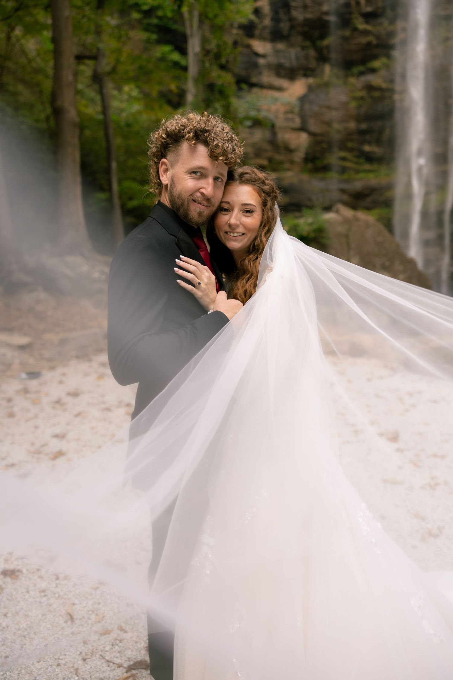 A romantic wedding photo captured at a misty waterfall with a flowing white veil creating an ethereal atmosphere.
