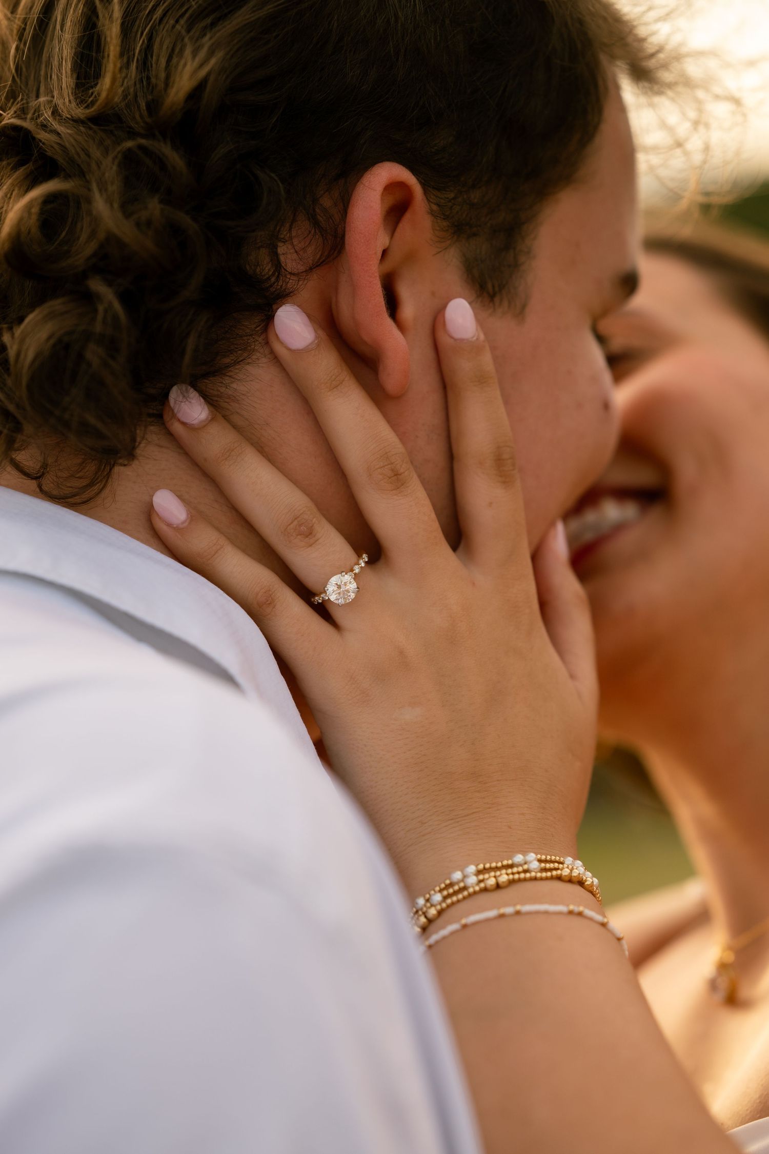 Close-up of a woman's hand with a diamond engagement ring and gold bracelets resting on a man's cheek as they kiss.