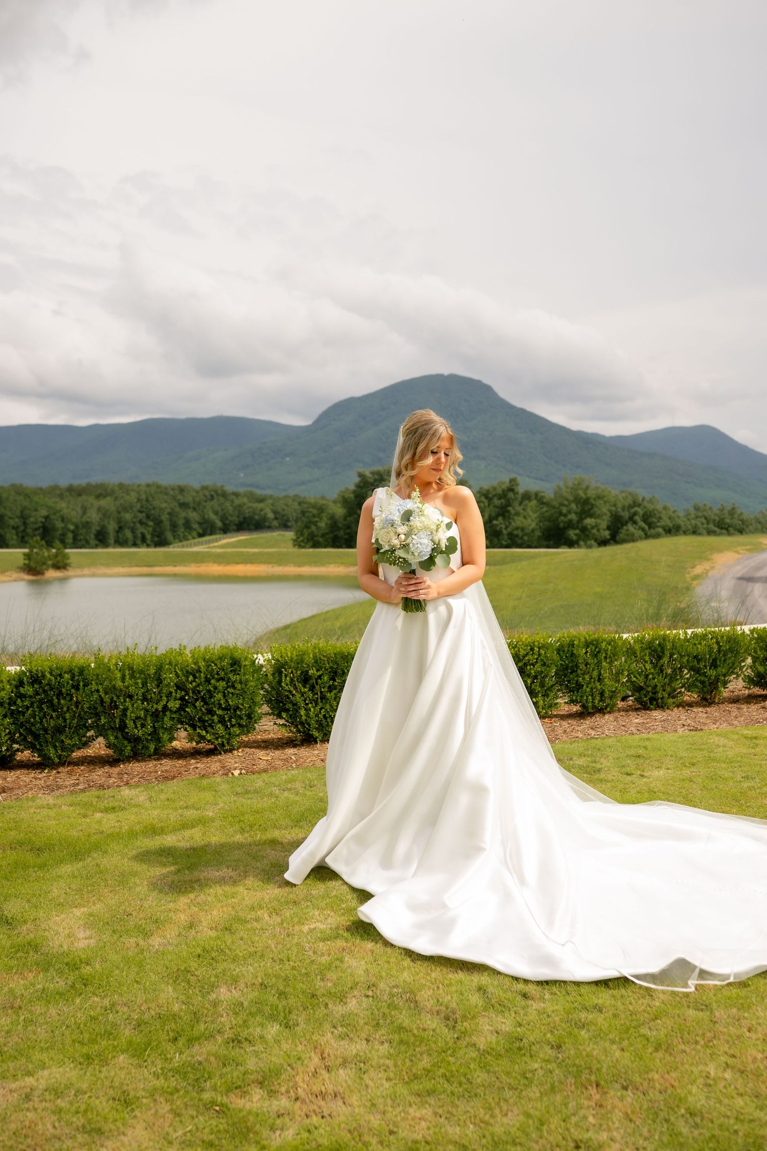 Bride in white gown with train holds bouquet, posing before mountain lake backdrop on lush green lawn.