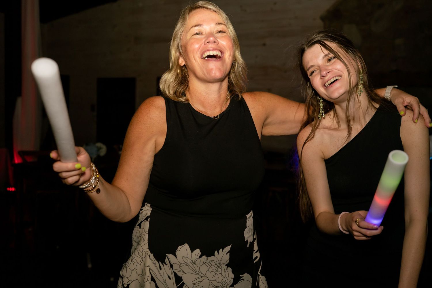Two women laughing and dancing at a party, holding glowing foam sticks, wearing elegant dresses at an evening event.