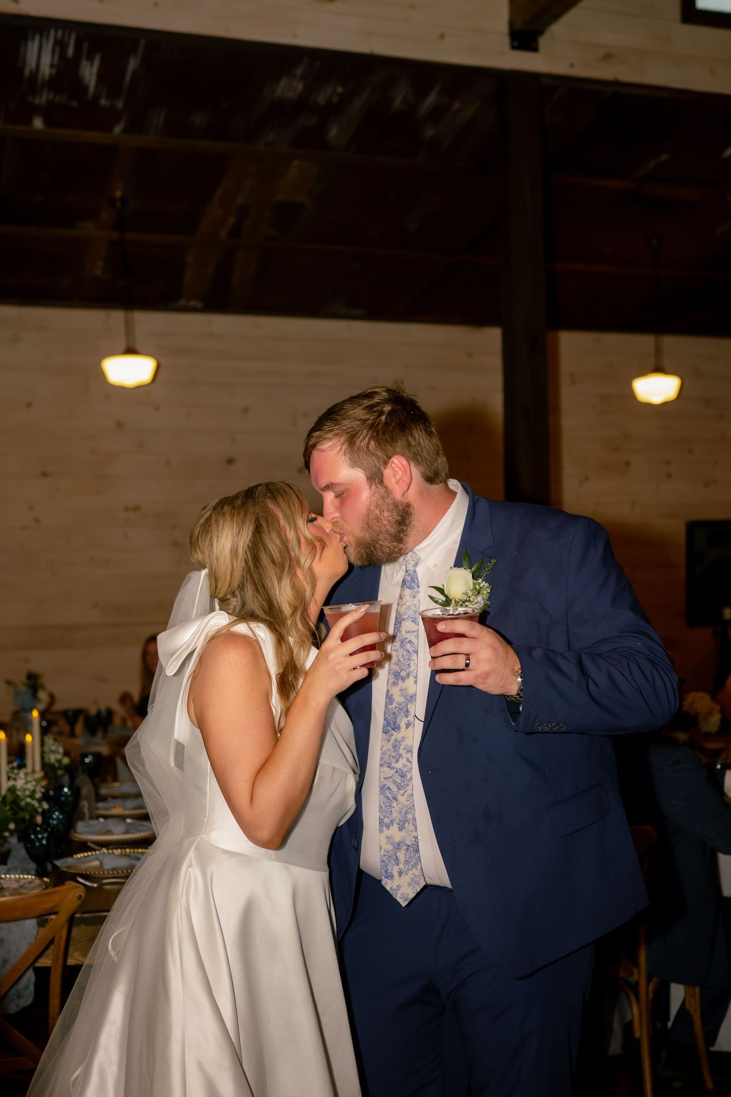 Bride and groom share a kiss while holding drinks at their rustic barn wedding reception.