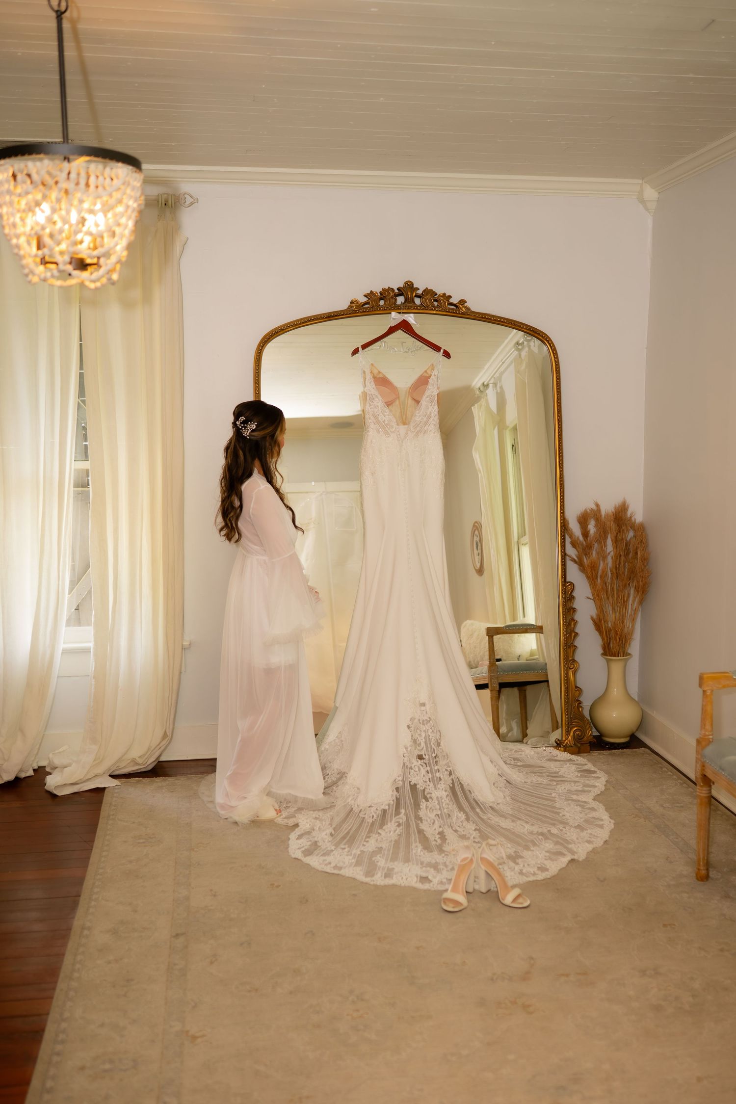Bride in pink robe admiring her elegant lace wedding gown hanging on a gold ornate mirror in a bright bridal suite.