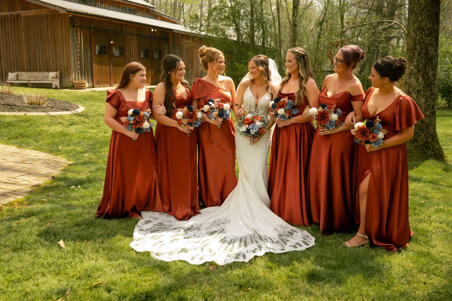 Bride in lace gown with bridesmaids in rust-colored dresses holding colorful bouquets on green lawn at rustic venue.