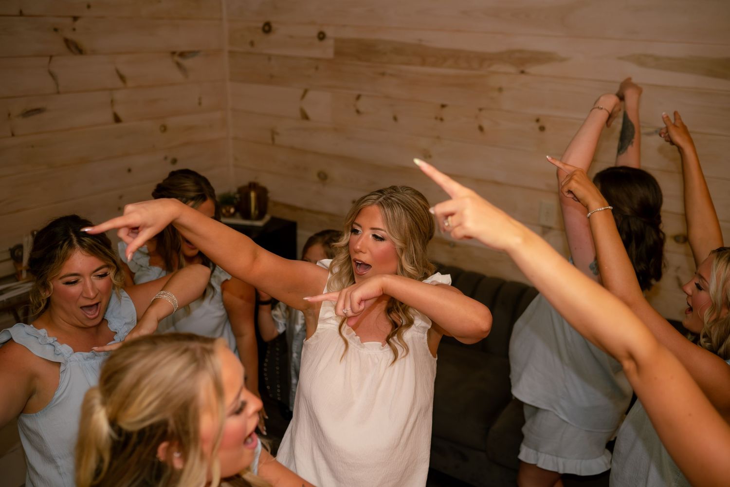 Bride in white dress dancing joyfully with bridesmaids in blue at a rustic wood-paneled wedding reception.