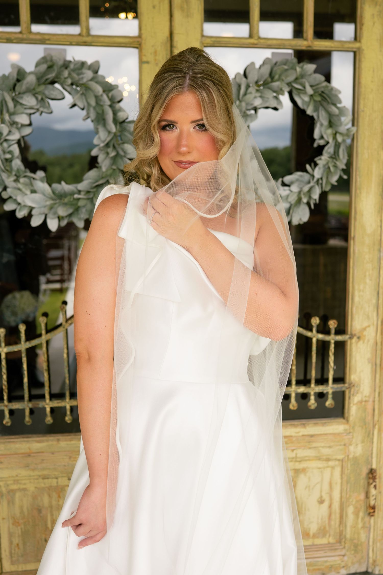Elegant bride in white satin gown and sheer veil poses before rustic doors with green leaf wreaths.