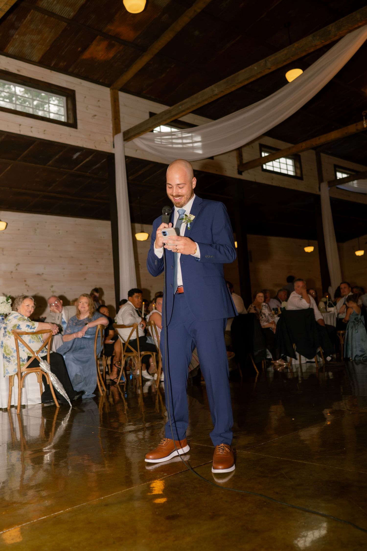 Groom in blue suit and brown shoes giving a speech with microphone at elegant rustic wedding reception venue.