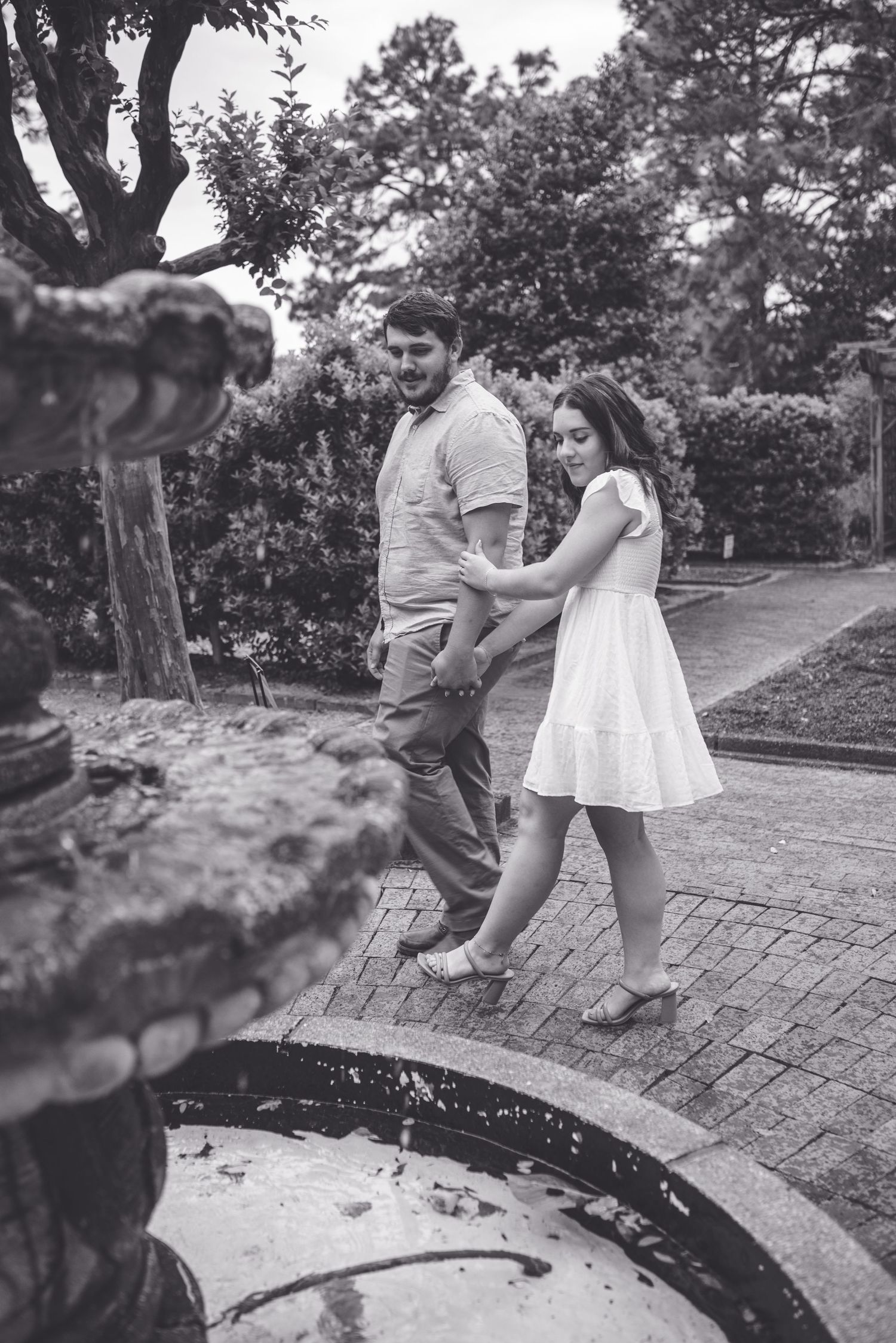 Black and white photo of a couple walking hand in hand near a stone fountain in a lush garden park.
