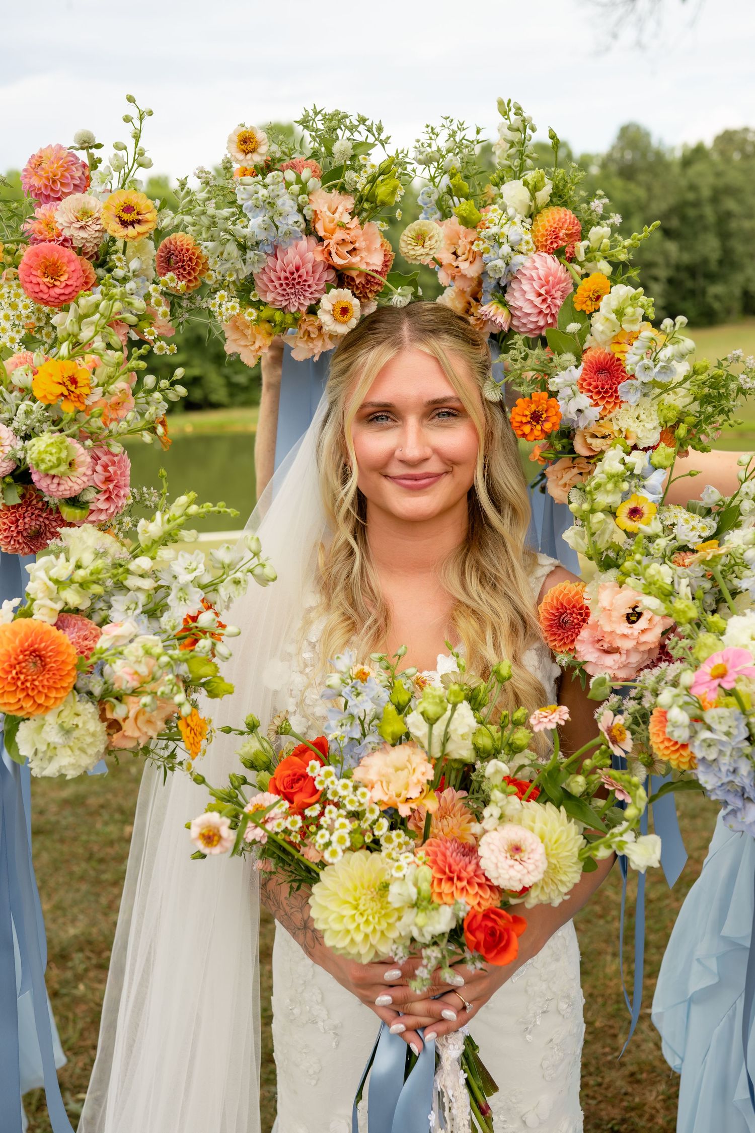 Smiling bride holds colorful bouquet framed by vibrant floral arch with dahlias and mixed blooms at outdoor wedding.