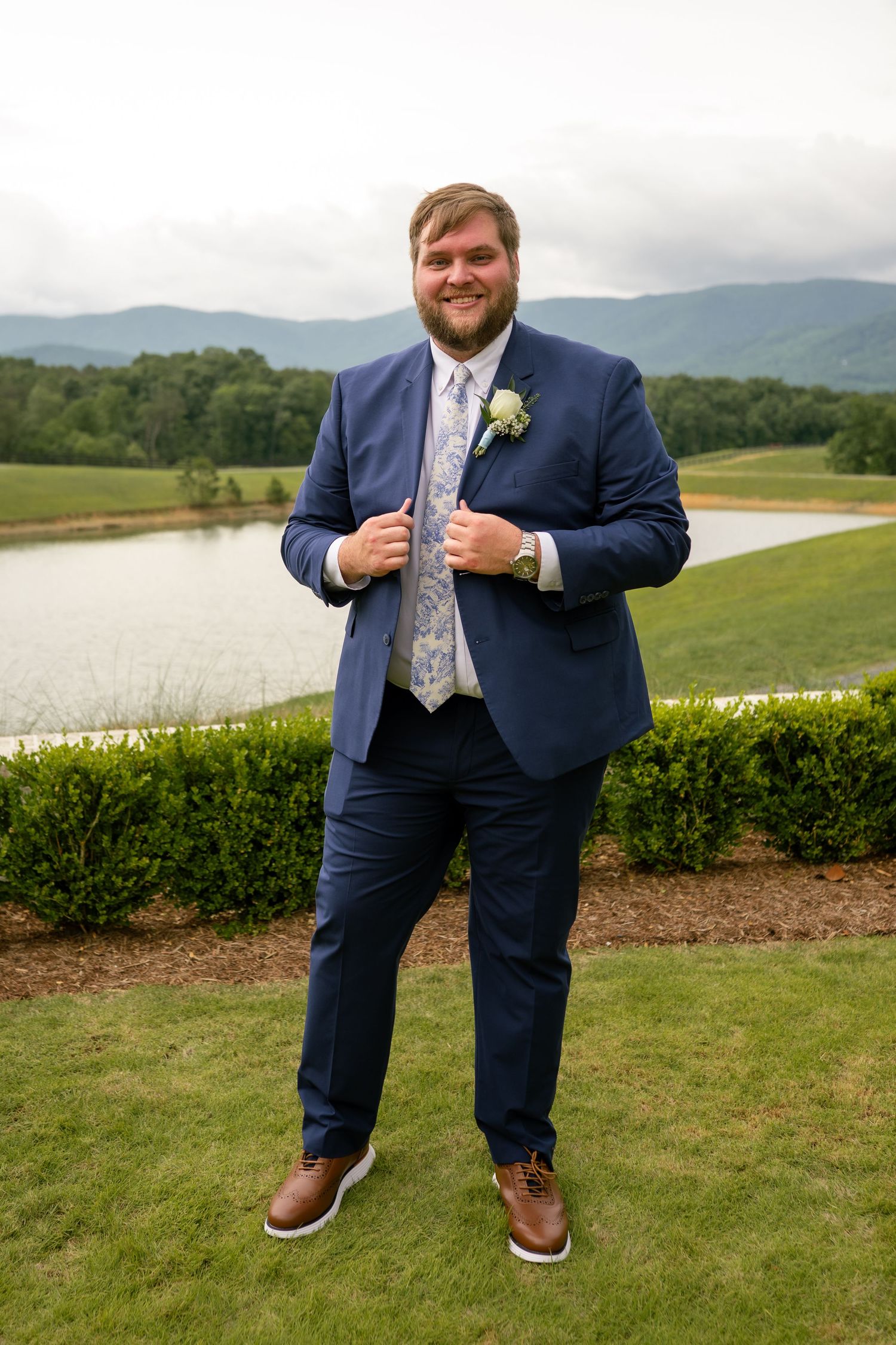 Groom in navy suit with floral tie and boutonniere posing outdoors near mountain lake.