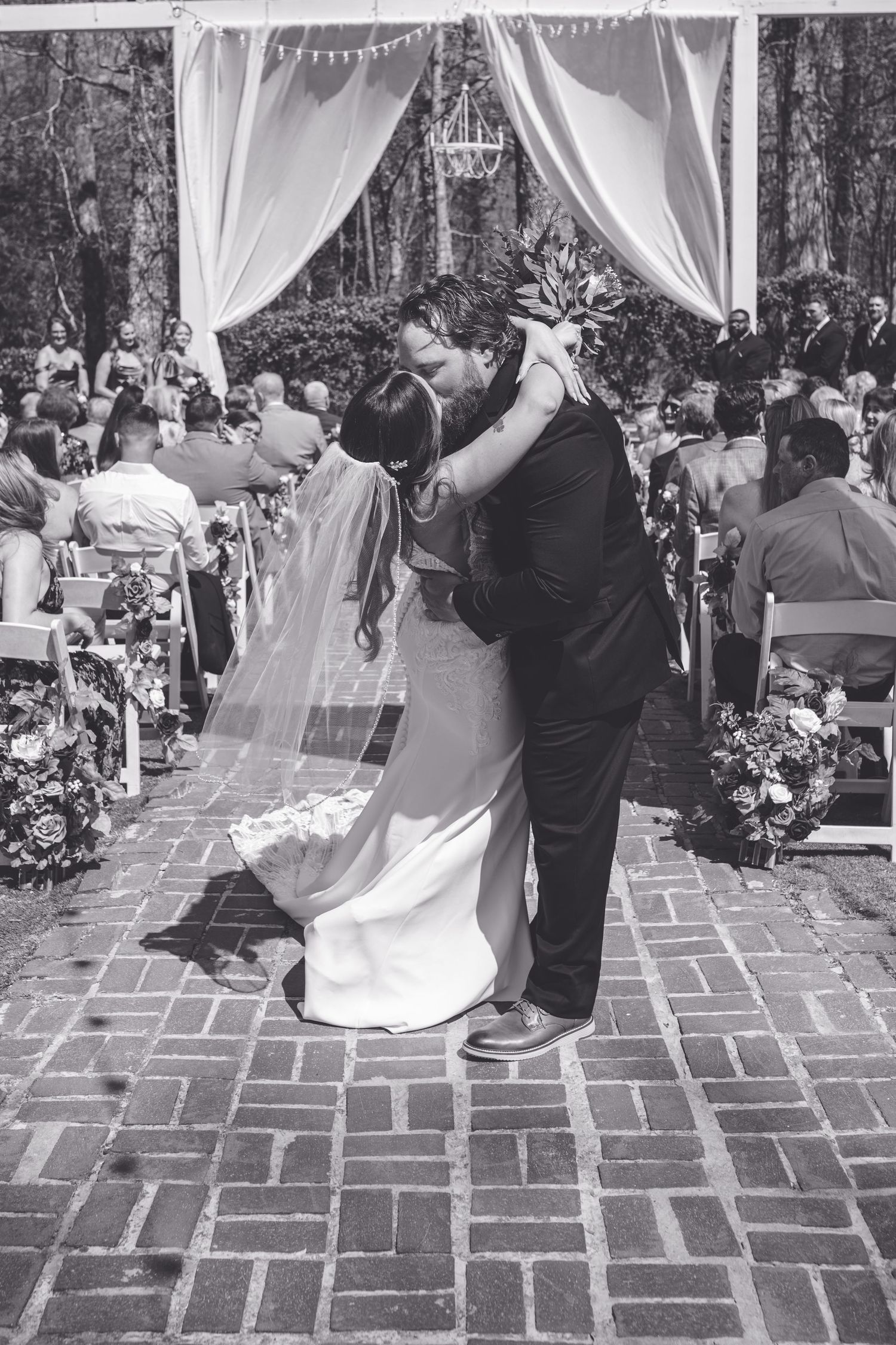 Bride and groom share first kiss at outdoor wedding ceremony under draped white canopy on brick path.