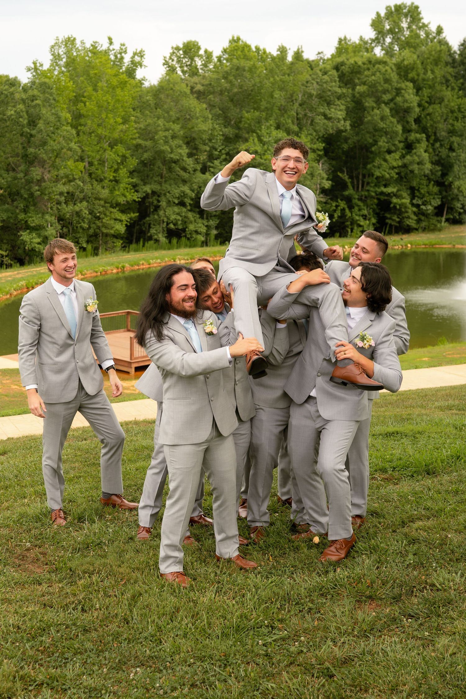 Groomsmen in gray suits lifting the groom outdoors near a pond at a wedding celebration.