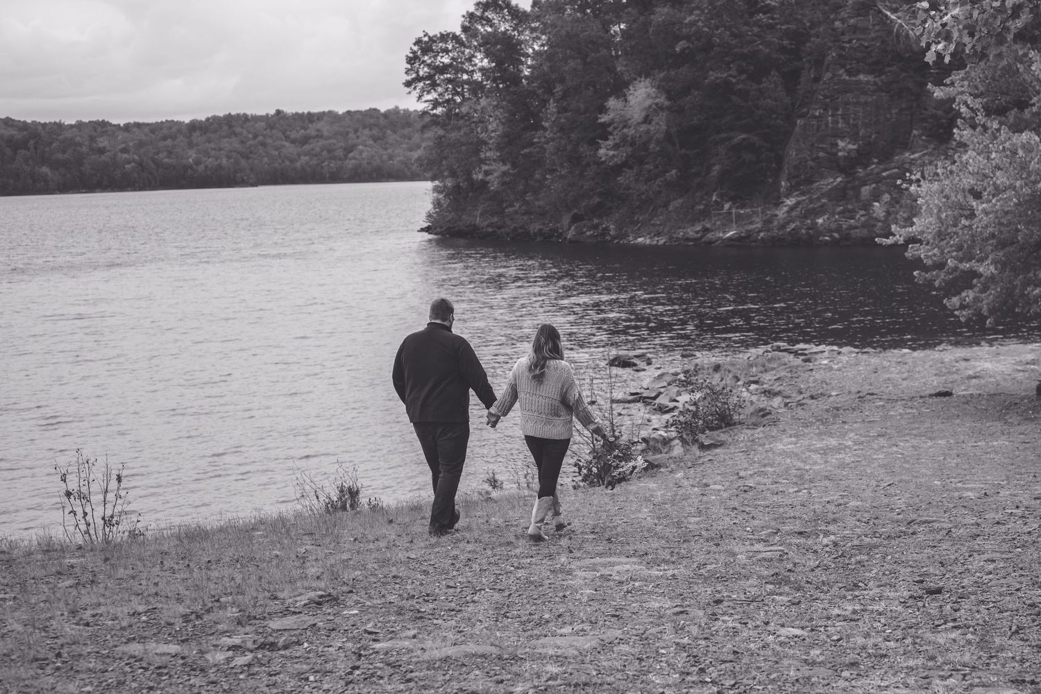 Couple holding hands walking along a rocky lakeside shore surrounded by lush trees in black and white.