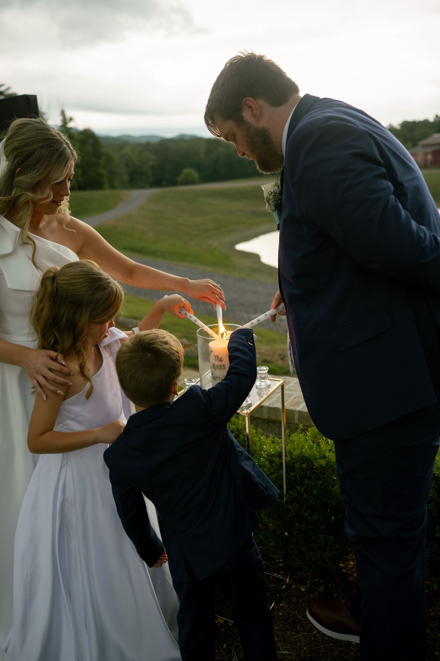Bride and groom light a unity candle with two children during an outdoor wedding ceremony.