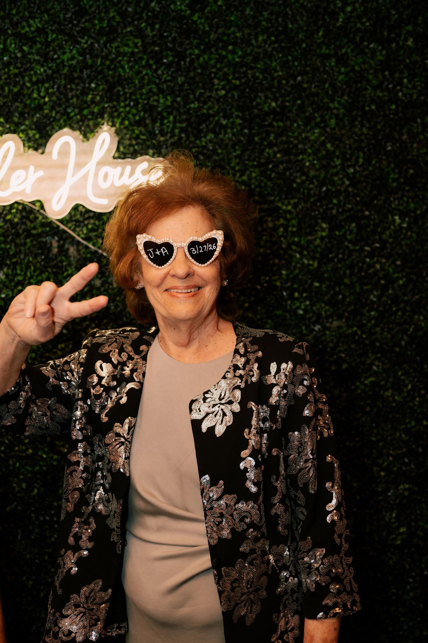 Elderly woman in sequined jacket poses with novelty heart-shaped glasses and peace sign at a party photo booth.
