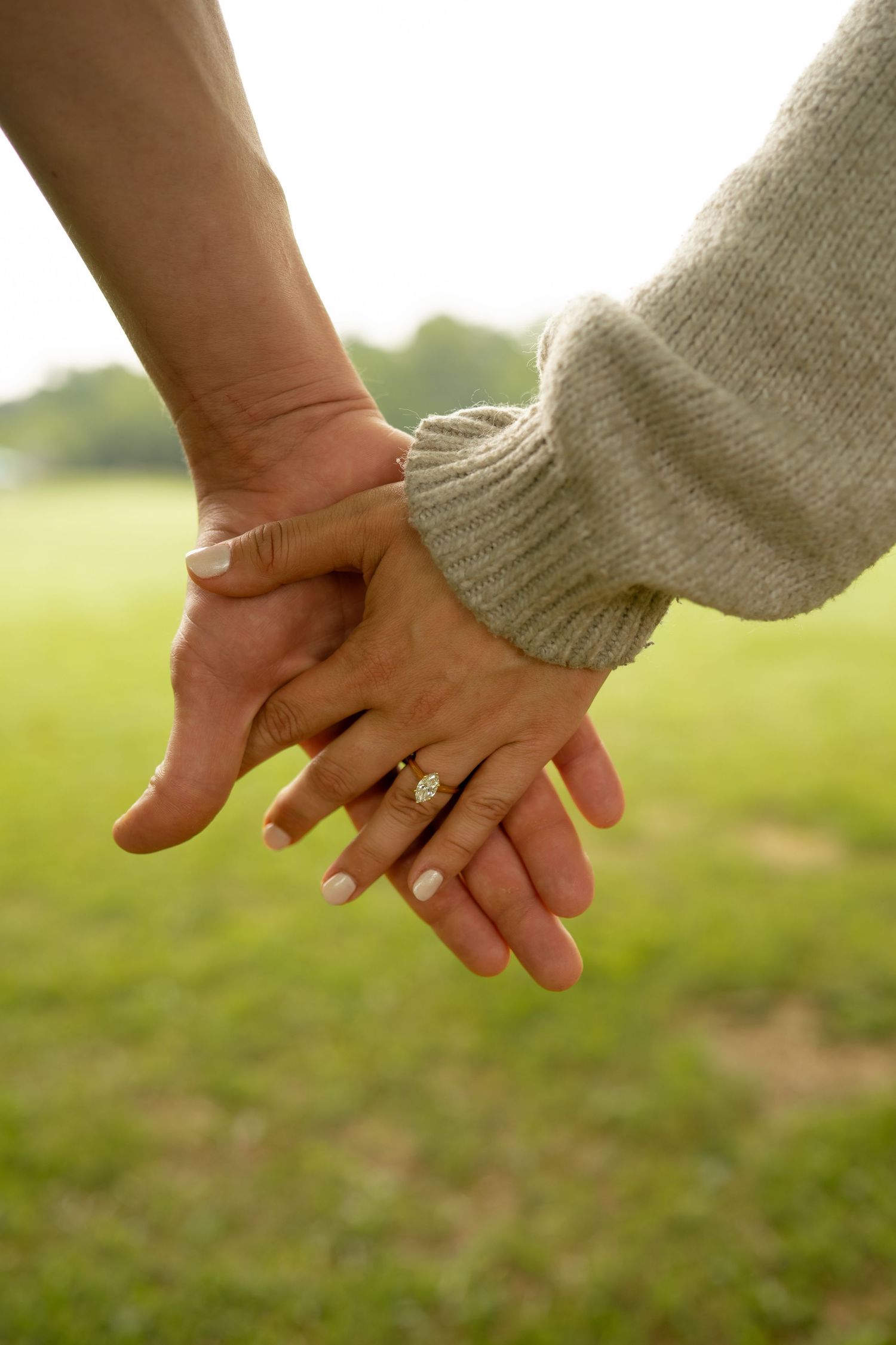 Close-up of a couple holding hands outdoors, woman wearing a diamond engagement ring, green field in background.