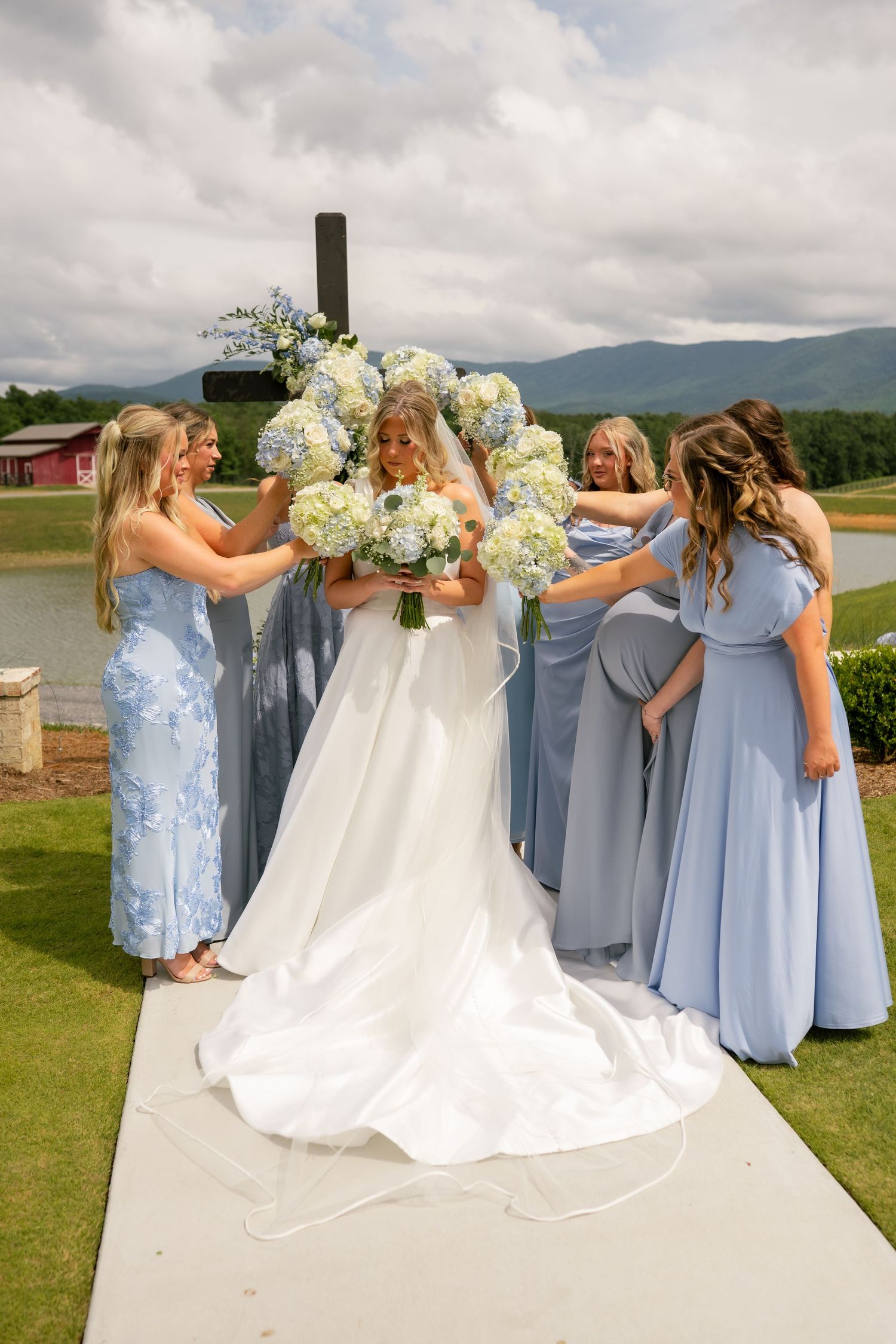 Bride in white gown surrounded by bridesmaids in blue dresses holding hydrangea bouquets outdoors with mountain backdrop.