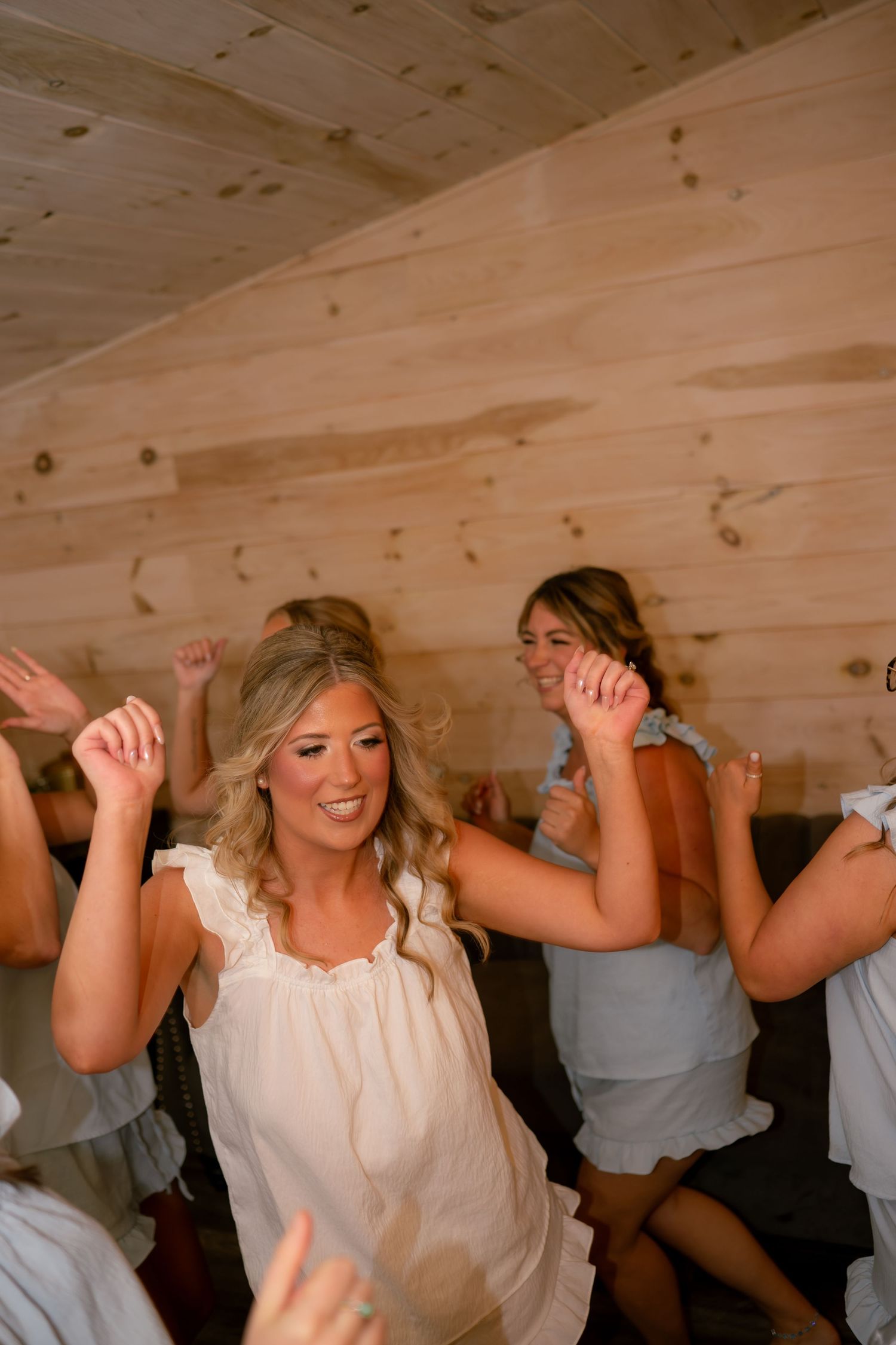 A blonde bride dances joyfully with guests in a rustic wood-paneled reception venue during her wedding celebration.