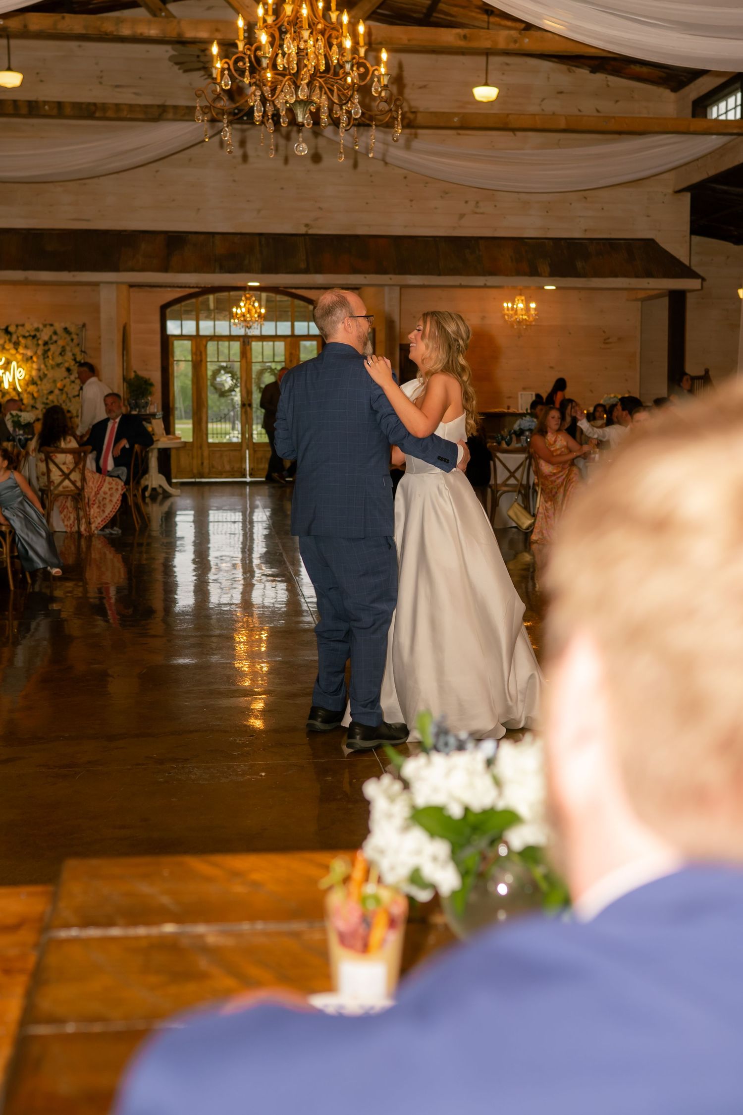 A bride in a champagne gown shares a touching father-daughter dance in a warmly lit rustic wedding reception venue.