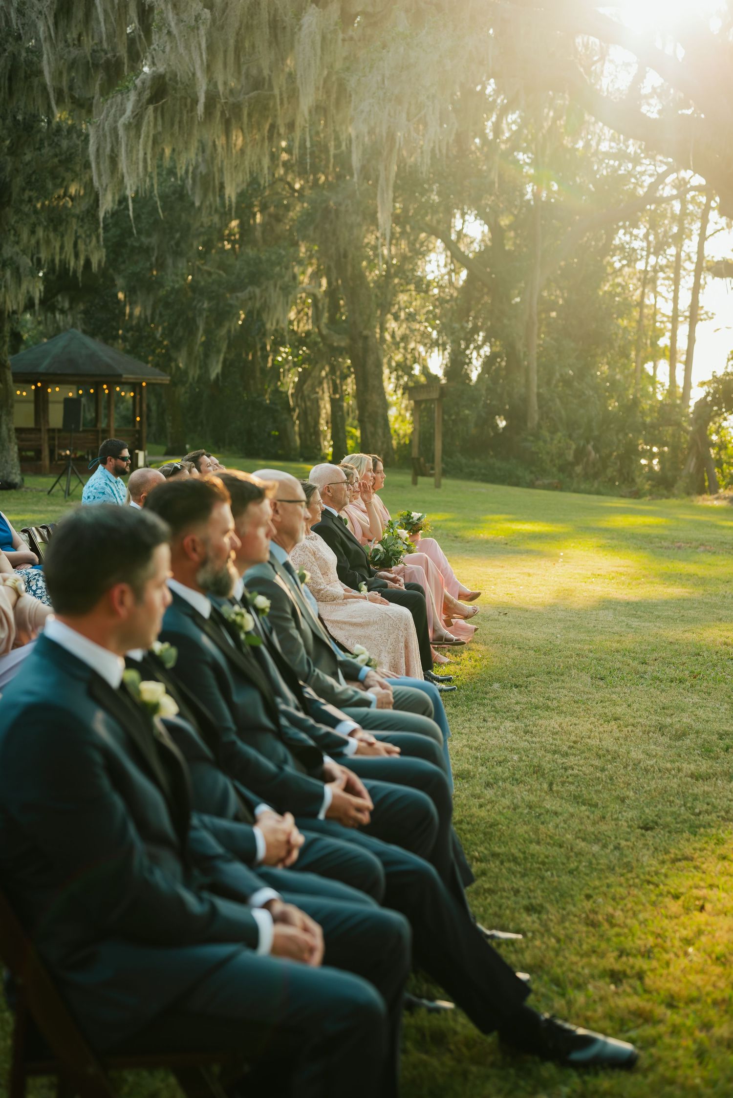Wedding guests seated in rows on a sunlit lawn with Spanish moss hanging from trees.