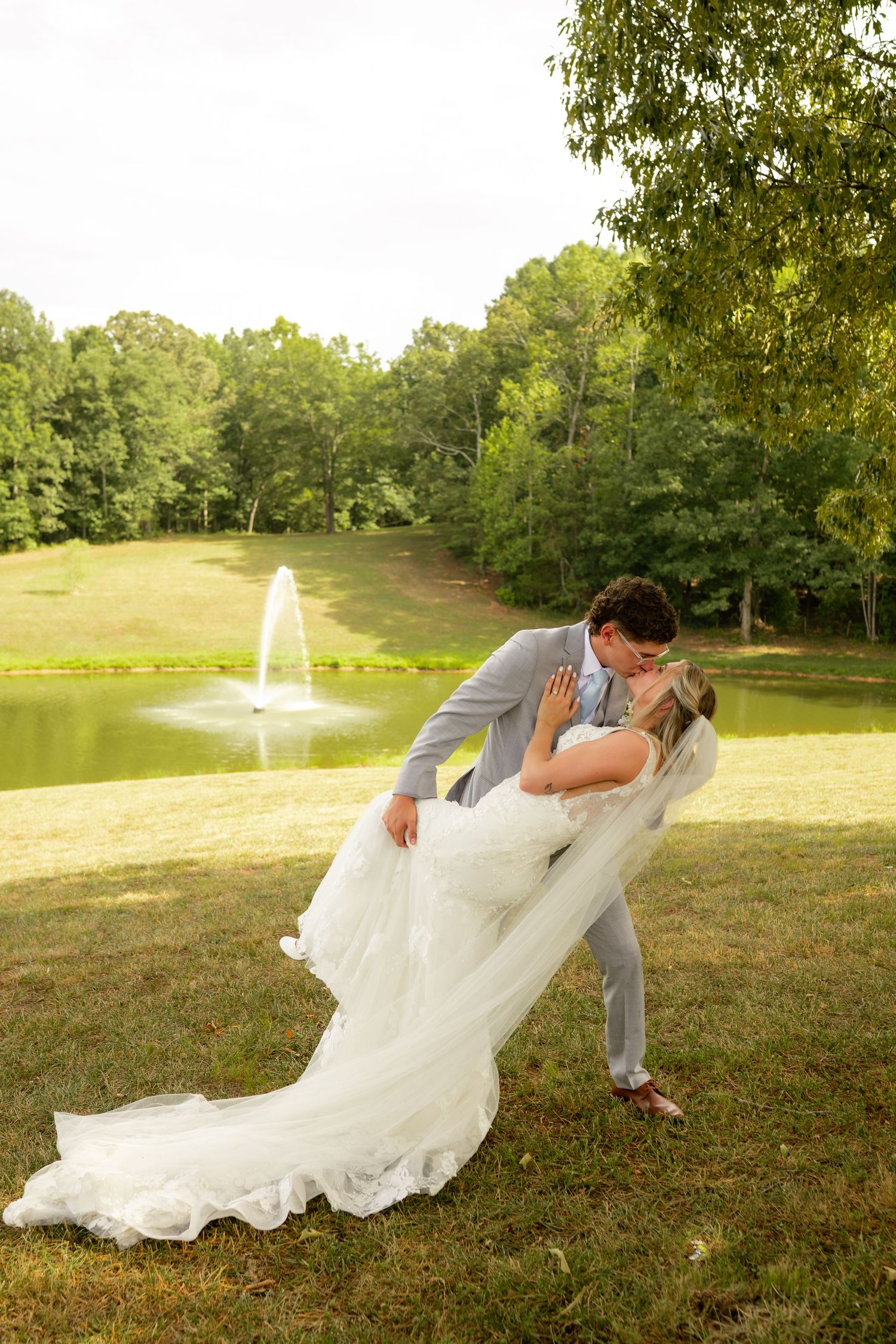 Groom dips bride in white gown for romantic kiss outdoors near pond with fountain surrounded by lush green trees.