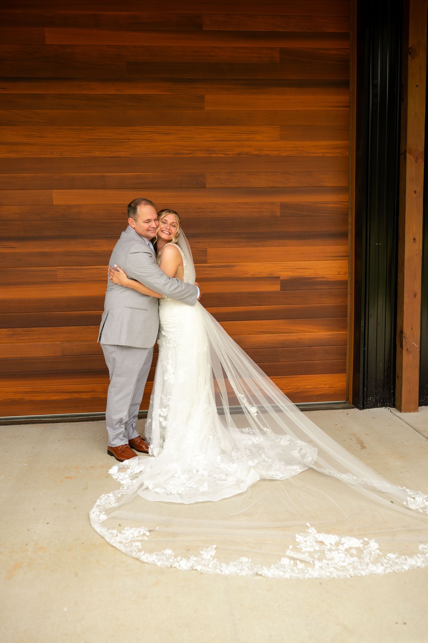 Bride and groom embracing in front of wooden wall, long lace veil trailing on ground.