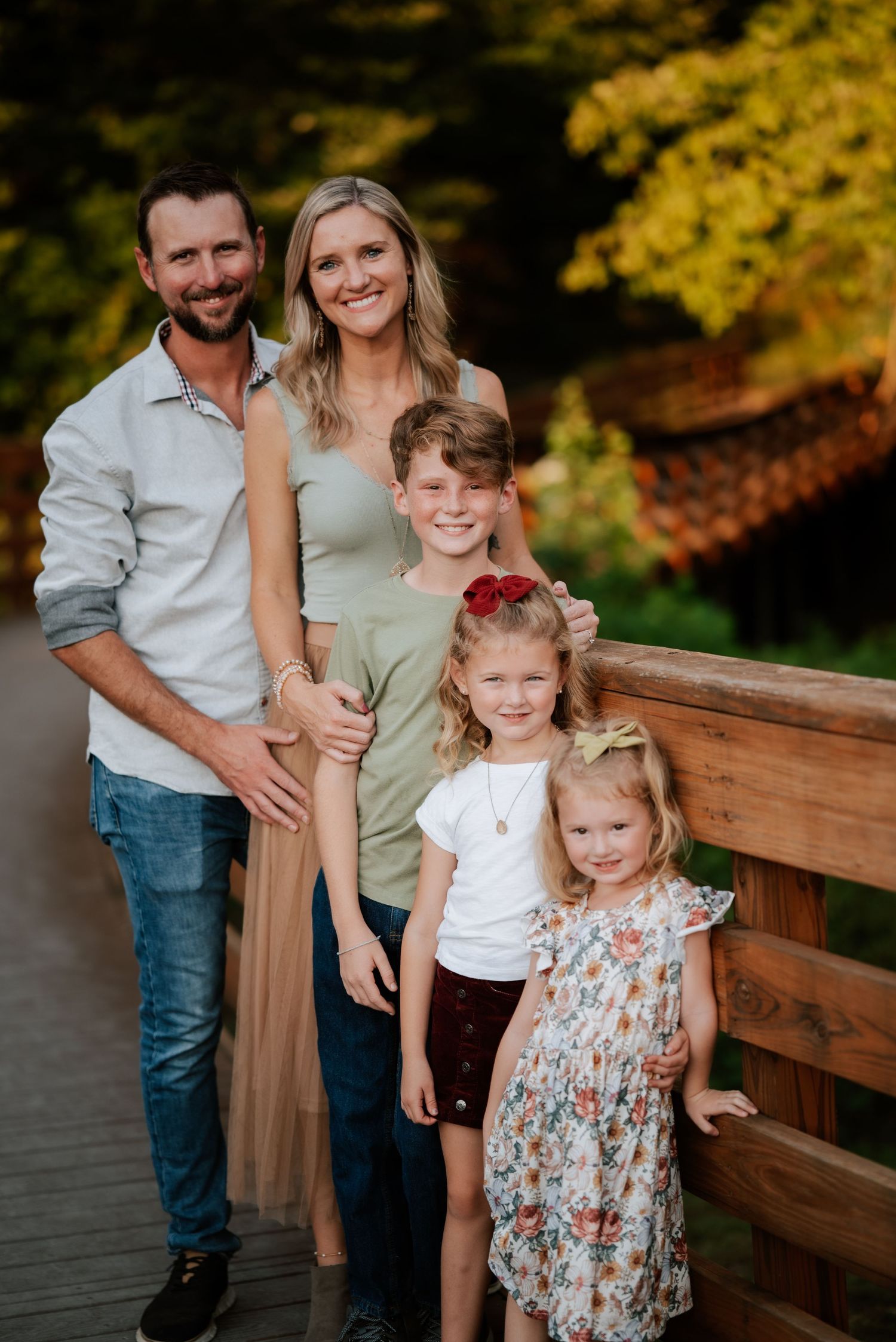 Family of five poses together on wooden bridge with autumn foliage in background.