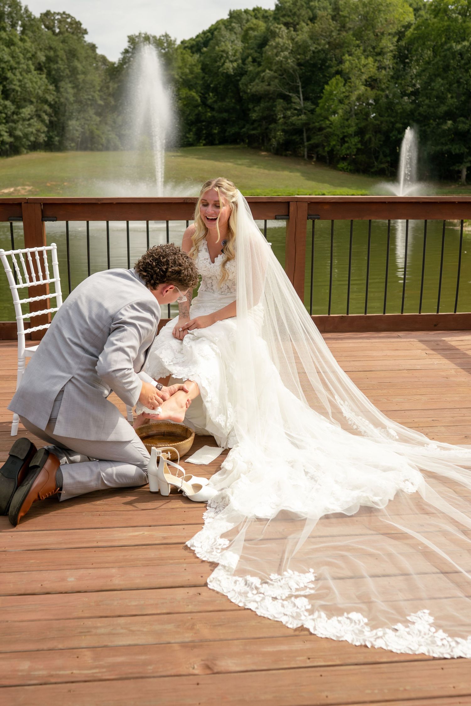 Groom kneels on wooden deck to put shoes on laughing bride in white gown with long veil near pond fountains.