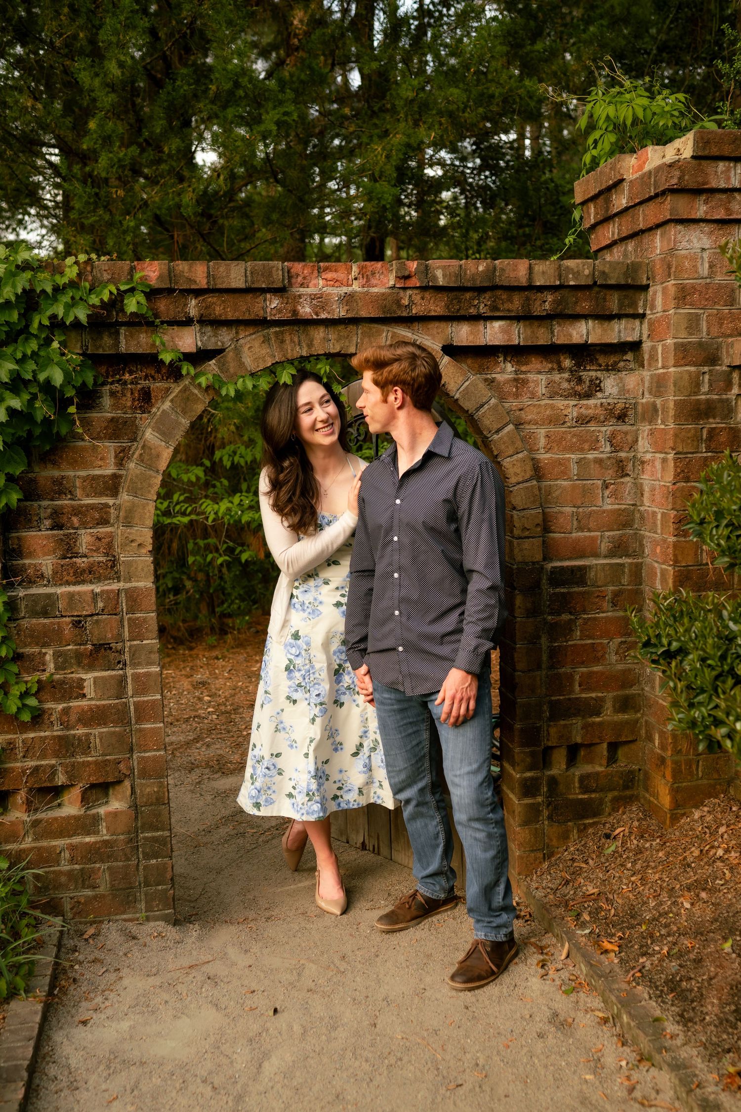 Couple smiling at each other under a brick archway surrounded by lush greenery in a romantic garden setting.