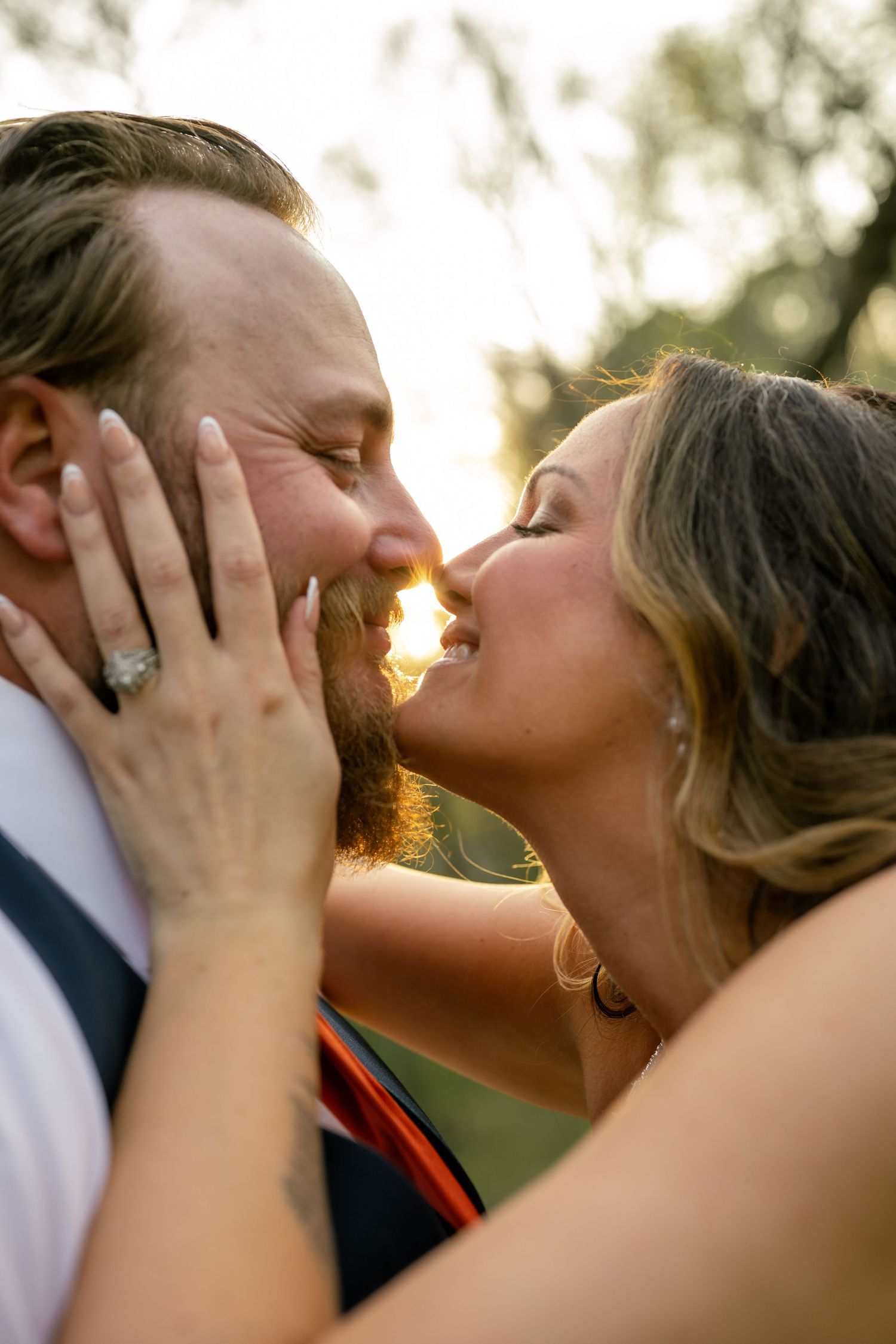 A couple shares a romantic kiss at golden hour, with the bride showcasing her engagement ring while holding her groom's face.