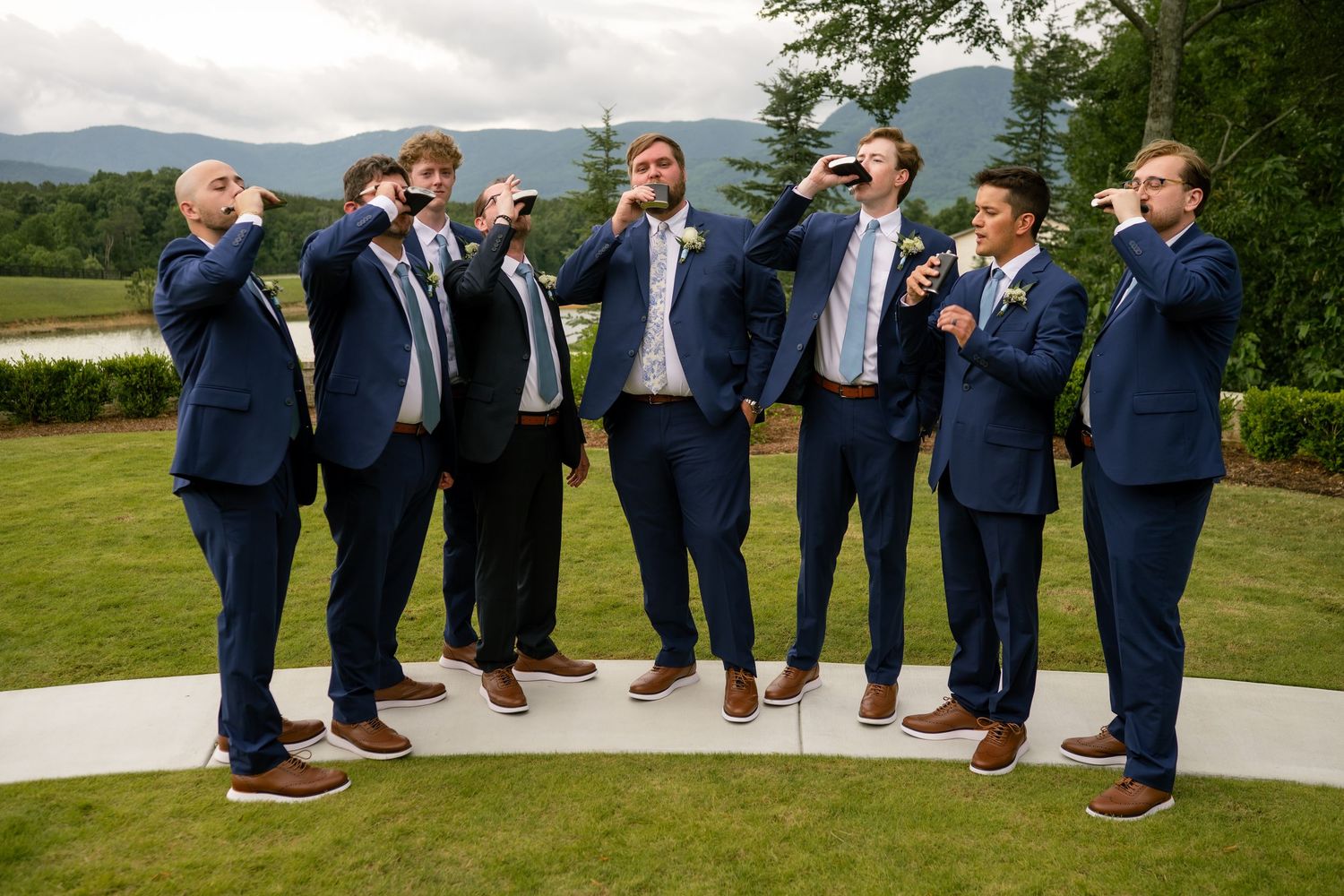 Seven groomsmen in navy suits toasting with drinks outdoors at a wedding venue with mountain views.